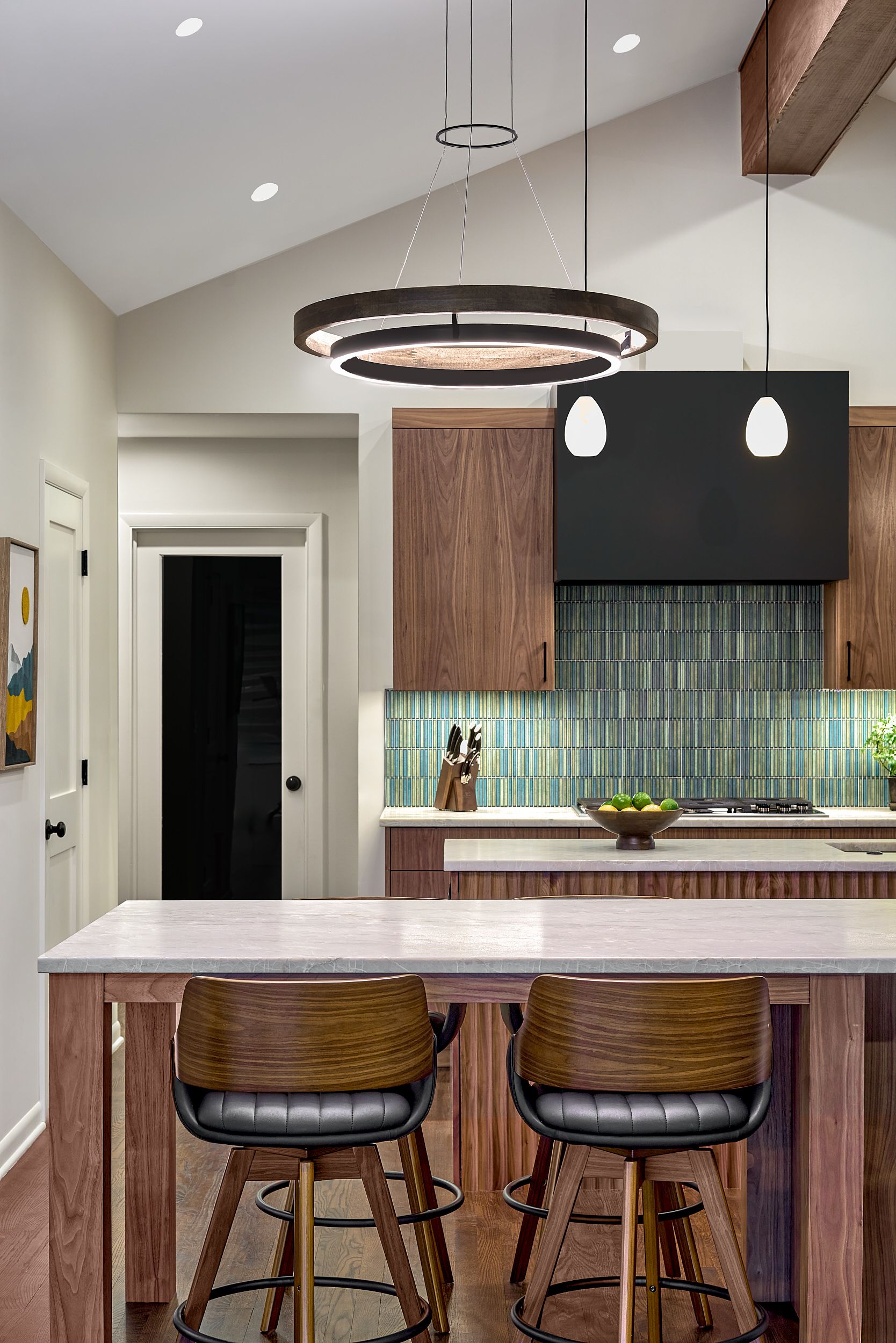 A contemporary kitchen features a large island with two bar stools, a wood-paneled kitchen with a blue and green tiled backsplash, and a black range hood.