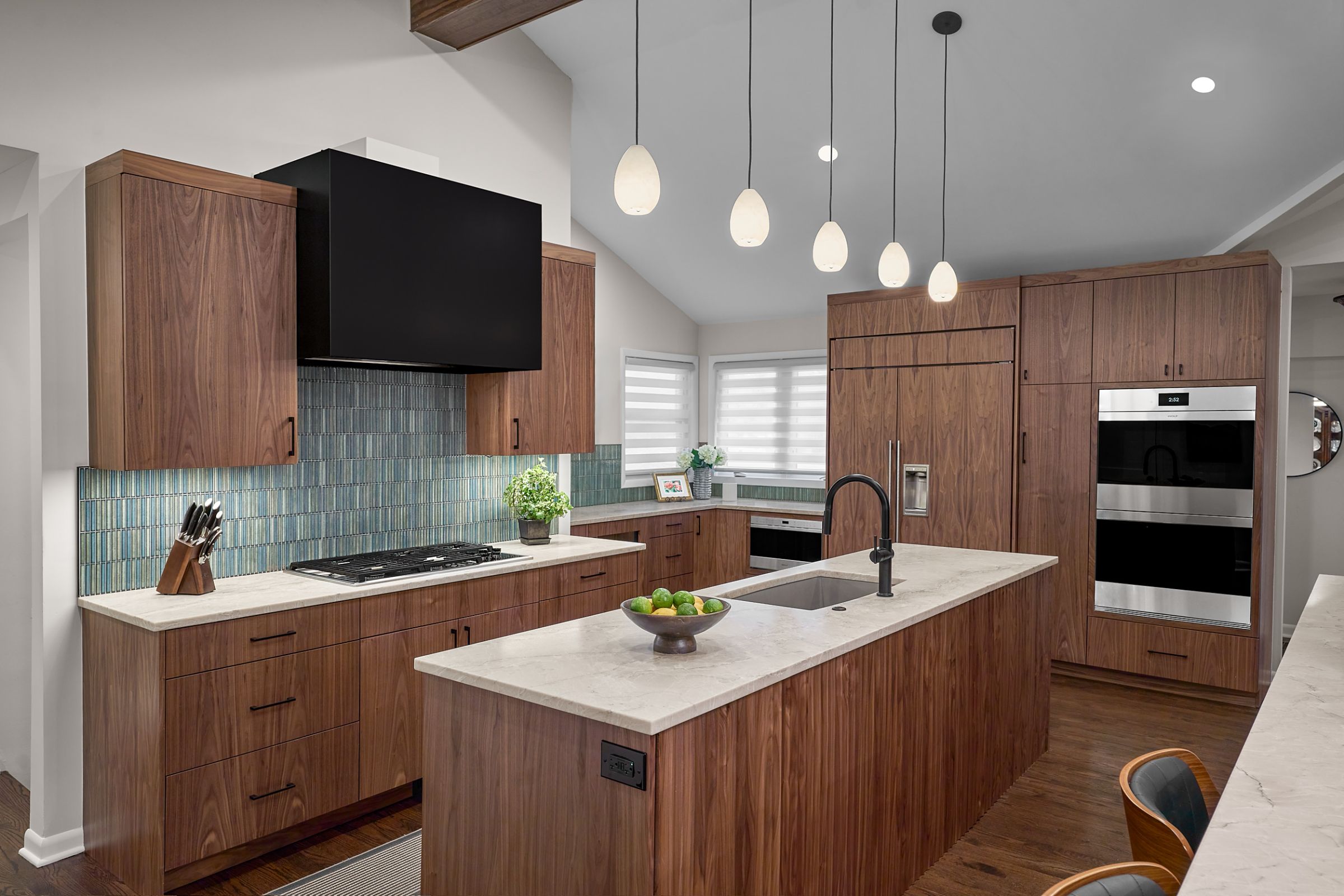 A kitchen with dark wood cabinetry, light countertops, a black range hood, and a kitchen island with a sink.