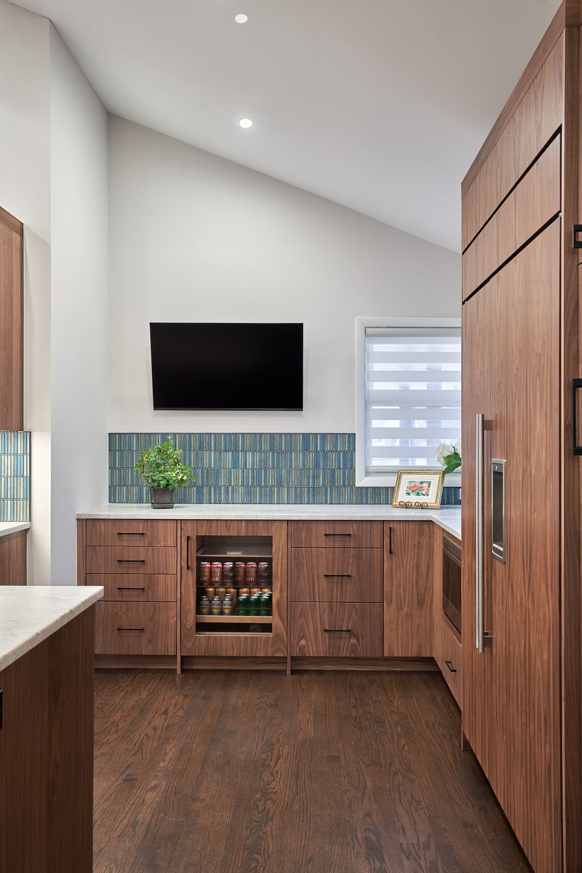A kitchen with dark wood cabinets, light countertops, and a blue and green tiled backsplash under a wall-mounted television. A beverage cooler is built into the cabinetry.