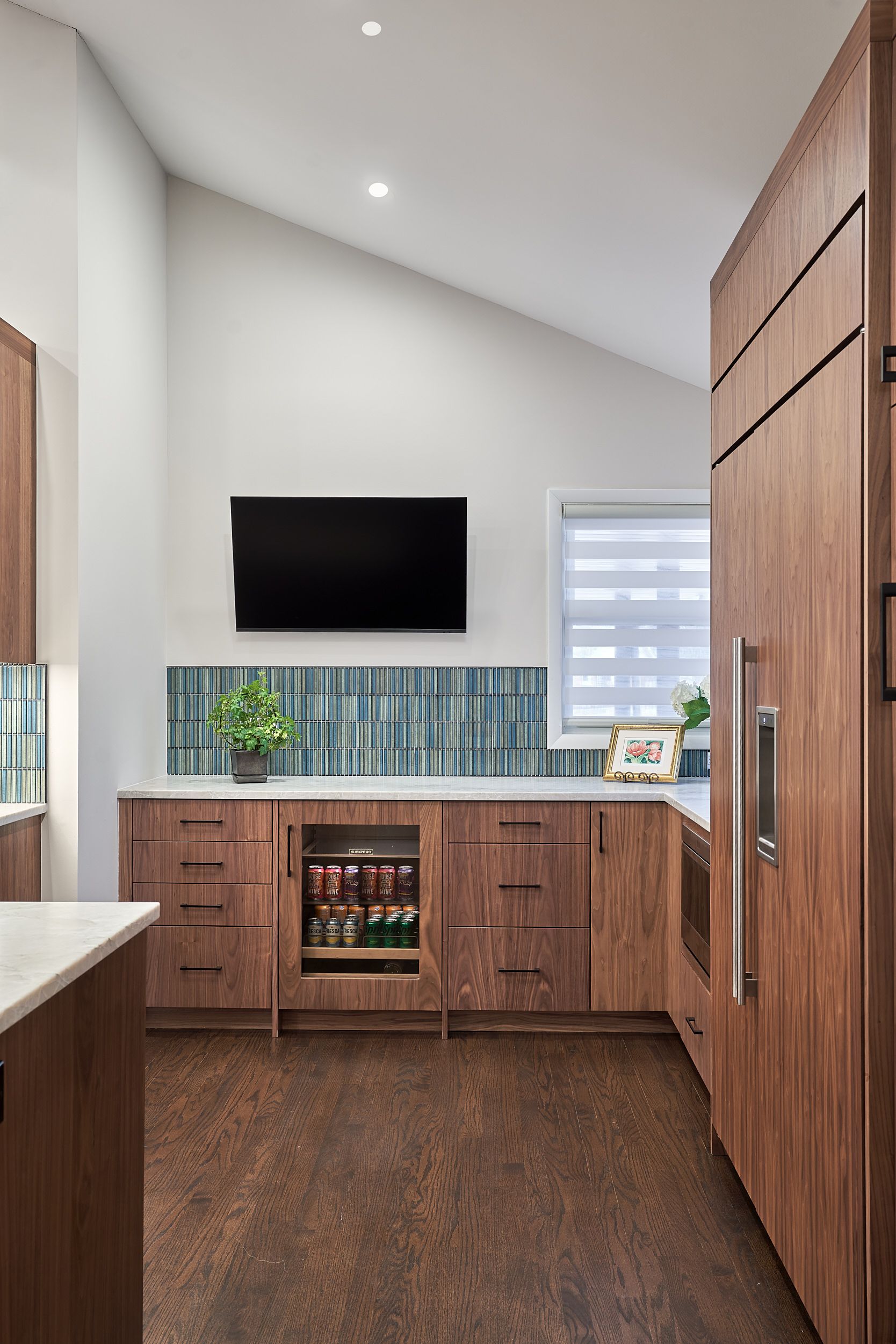 A kitchen with dark wood cabinets, light countertops, and a blue and green tiled backsplash under a wall-mounted television. A beverage cooler is built into the cabinetry.