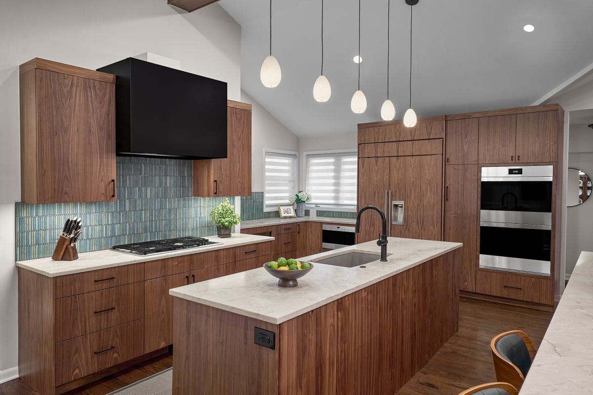 A kitchen with dark wood cabinetry, a light countertop, a blue tiled backsplash, and an island with a sink and pendant lights overhead.