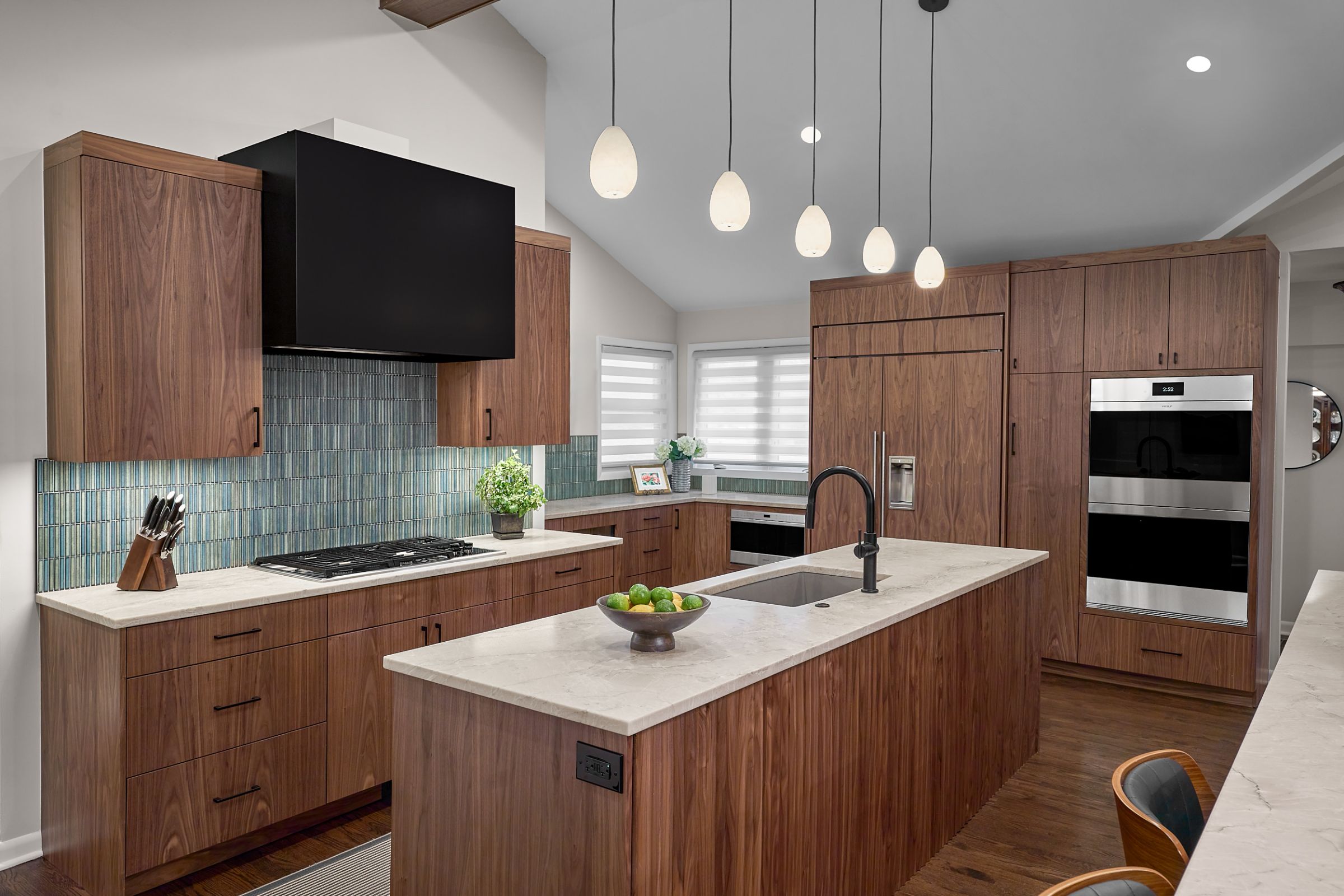 A kitchen with dark wood cabinetry, a light countertop, a blue tiled backsplash, and an island with a sink and pendant lights overhead.
