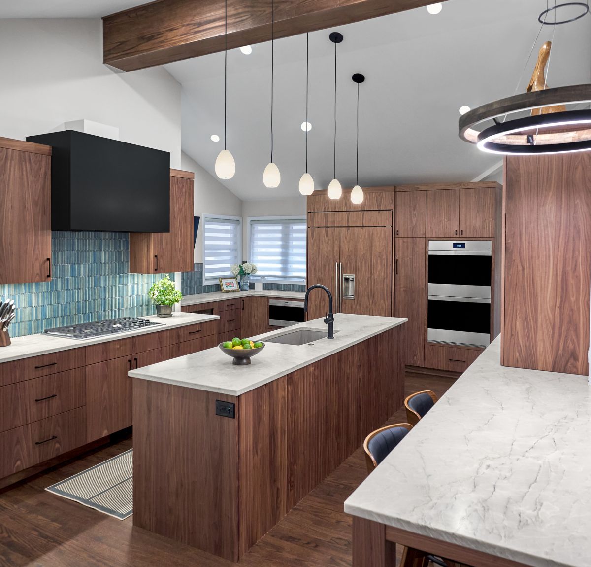 An open-concept kitchen features dark wood cabinetry, a large island with a sink, and light countertops, with a black range hood and blue tile backsplash.