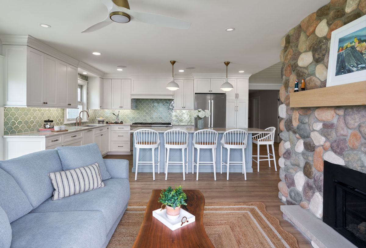 An open-concept living room and kitchen features a light blue sofa, a wood coffee table, a kitchen island with bar stools, and a stone fireplace.