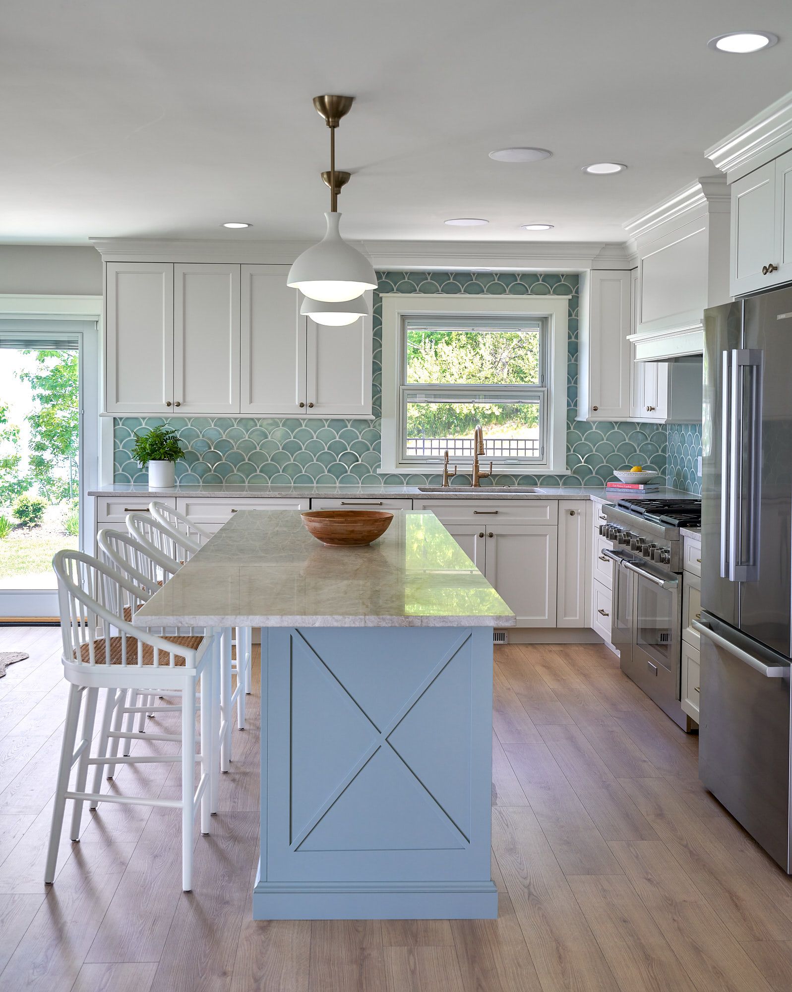 A bright kitchen features white cabinets, a blue island with seating, a teal tile backsplash, and stainless steel appliances.