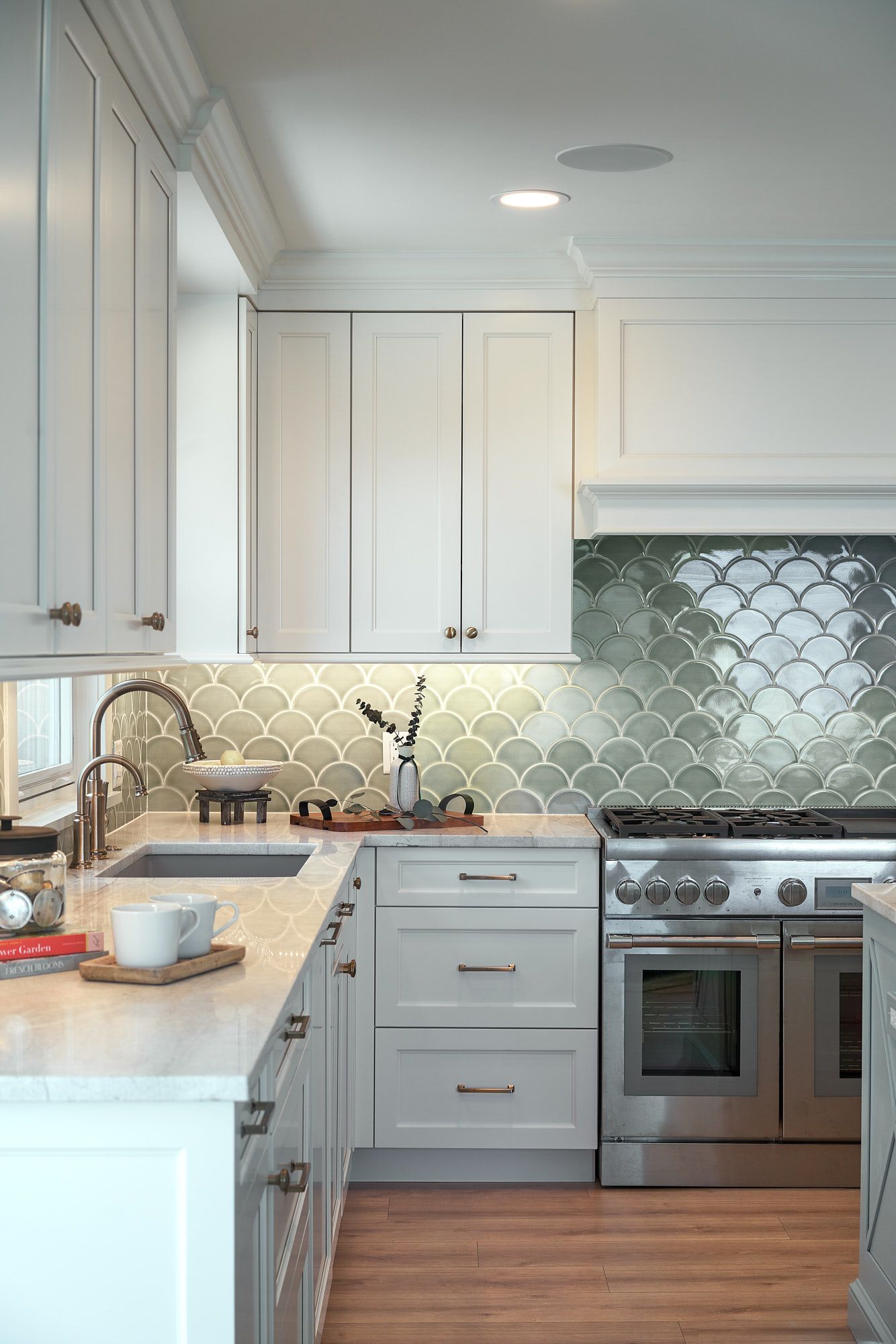 A kitchen features white upper and lower cabinets, a light-colored countertop, a sink, a stainless steel range, and a green scalloped tile backsplash.