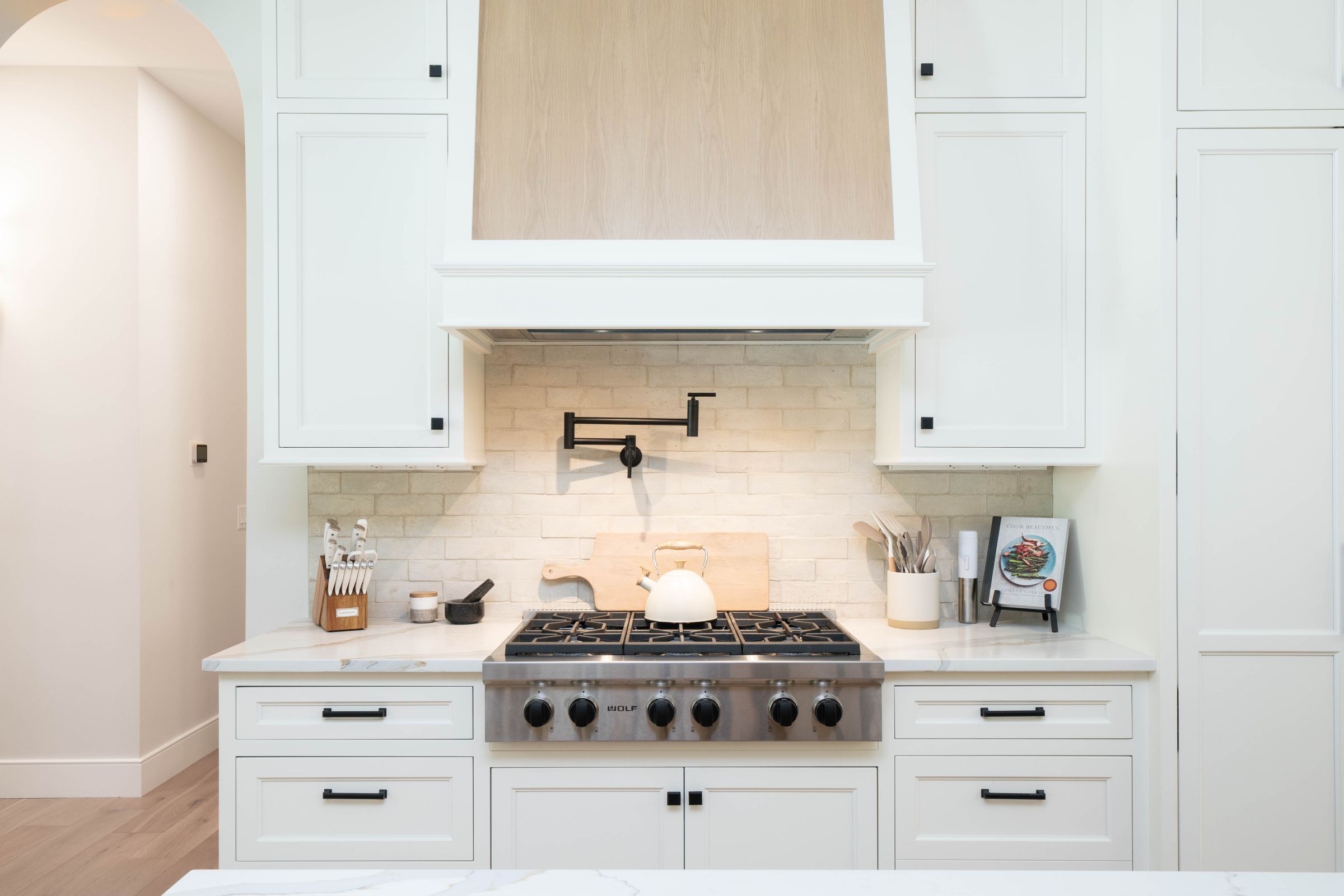 A white kitchen features a stainless steel range below a pot filler and a white brick backsplash, with white cabinets and light wood flooring.
