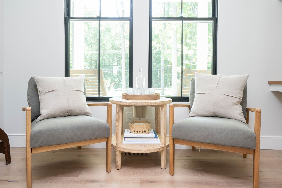 Two armchairs with light gray cushions and light wood frames flank a round wooden side table in front of two large windows.