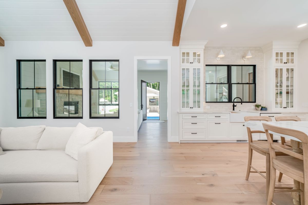 A bright open-plan living room and kitchen with white walls, light wood floors, a white sofa, and a kitchen island with chairs. Black-framed windows are visible throughout the space, and a doorway leads to an outdoor area with a pool.