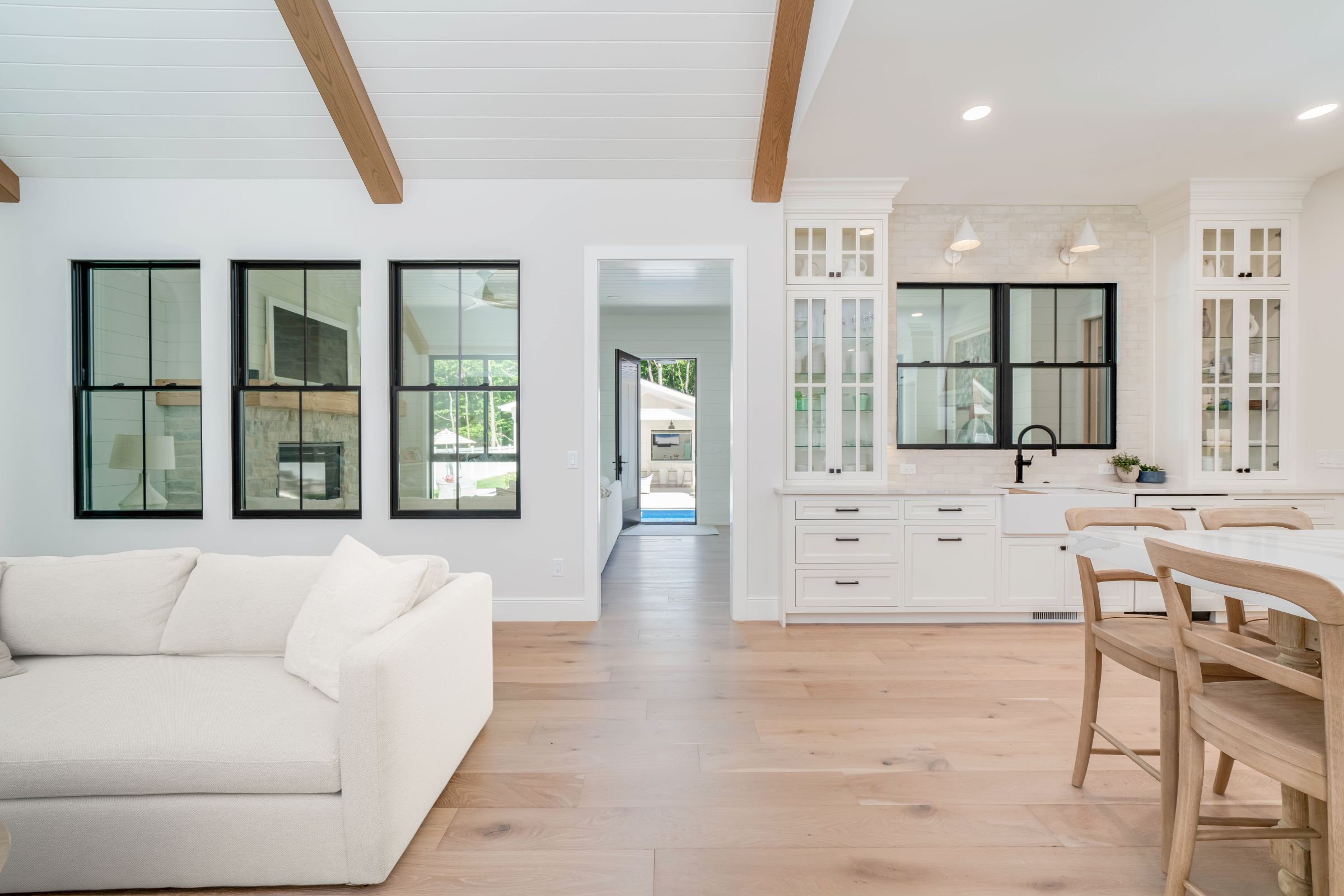 A bright open-plan living room and kitchen with white walls, light wood floors, a white sofa, and a kitchen island with chairs. Black-framed windows are visible throughout the space, and a doorway leads to an outdoor area with a pool.