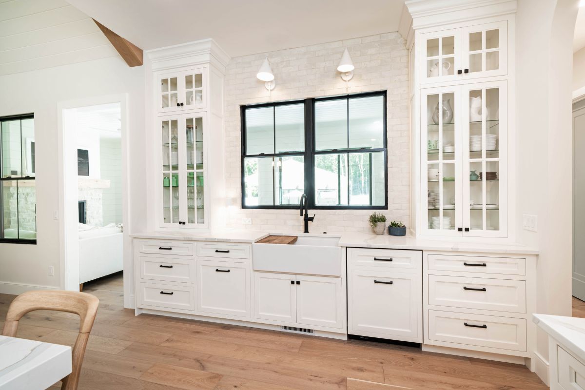 A white modern farmhouse kitchen features white cabinetry, a white brick backsplash, and a large black-framed window above a farmhouse sink.