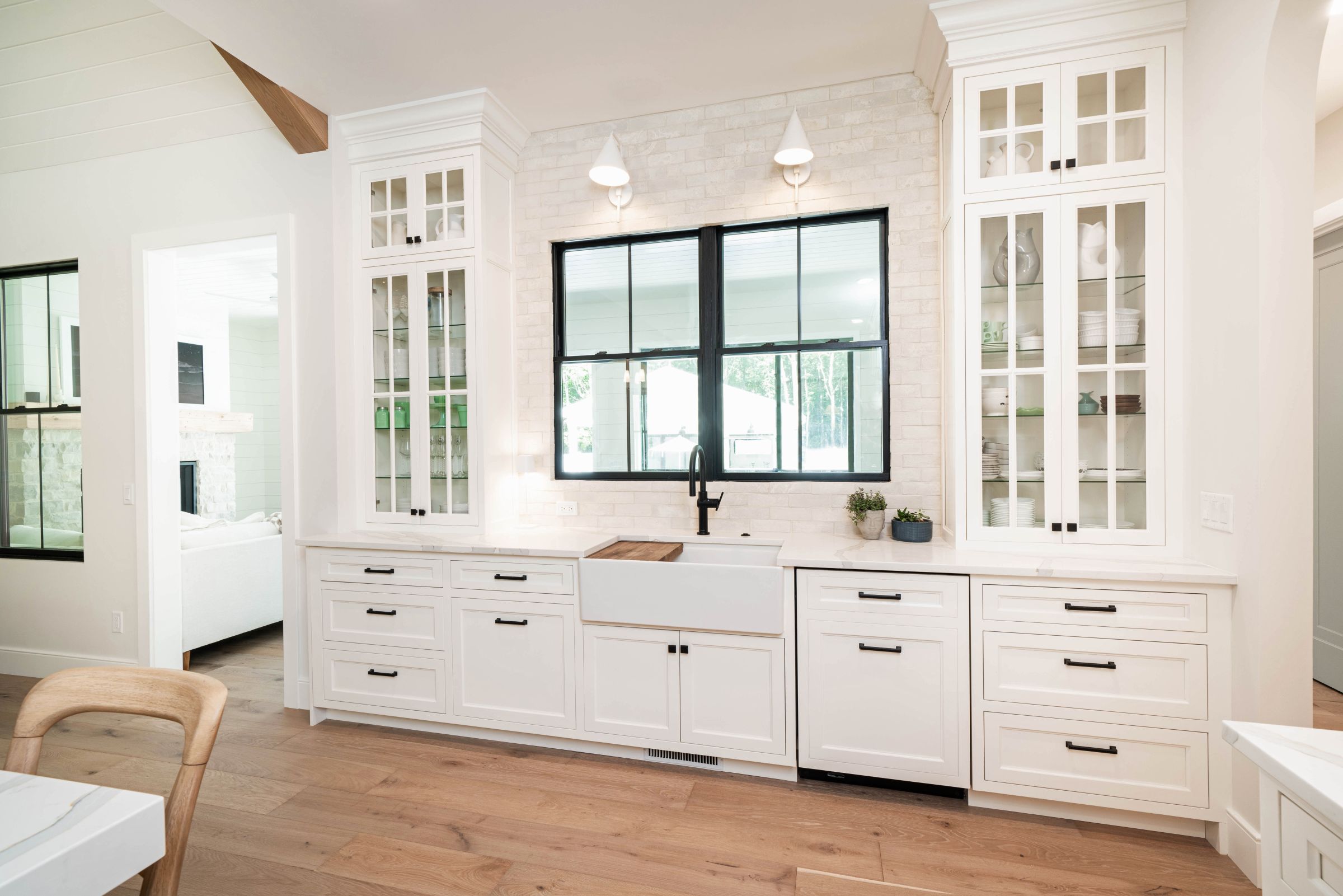 A white modern farmhouse kitchen features white cabinetry, a white brick backsplash, and a large black-framed window above a farmhouse sink.