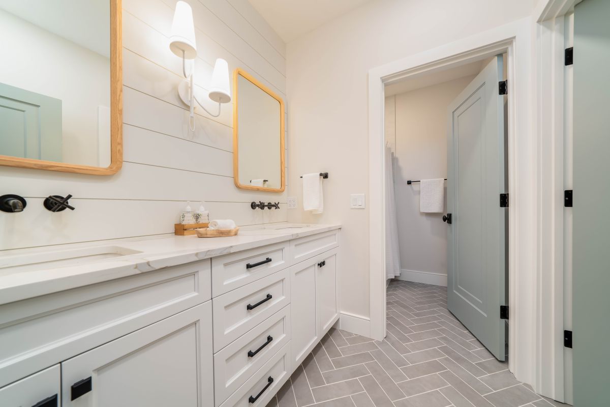 A modern farmhouse bathroom features a double vanity with white cabinets, light gray herringbone floor tiles, and two mirrors with sconces.