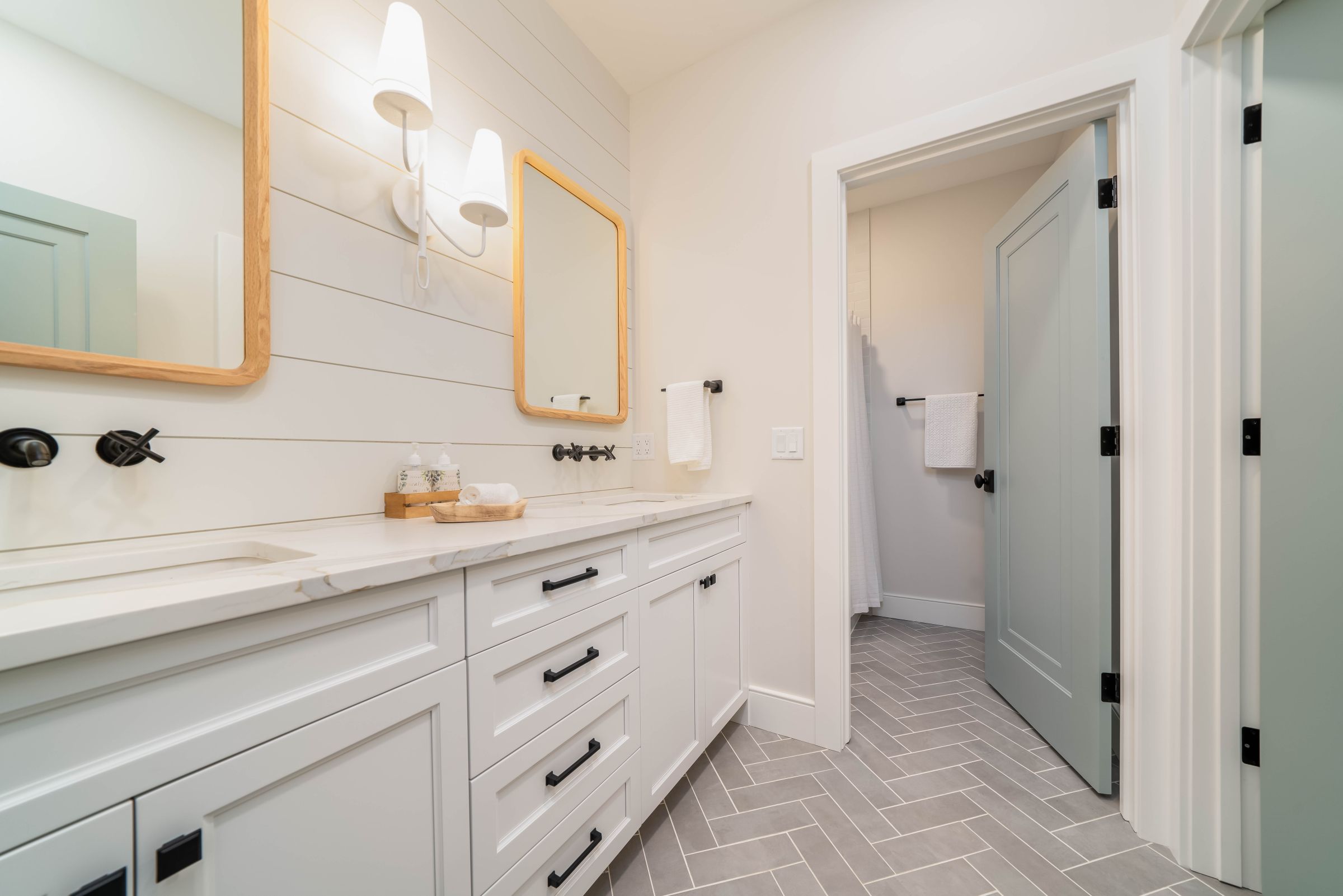 A modern farmhouse bathroom features a double vanity with white cabinets, light gray herringbone floor tiles, and two mirrors with sconces.