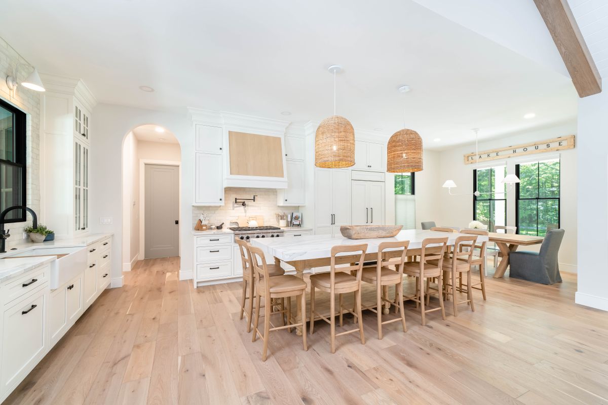 A bright kitchen with white cabinets, light wood floors, a large island with seating, and a dining area visible through large windows.