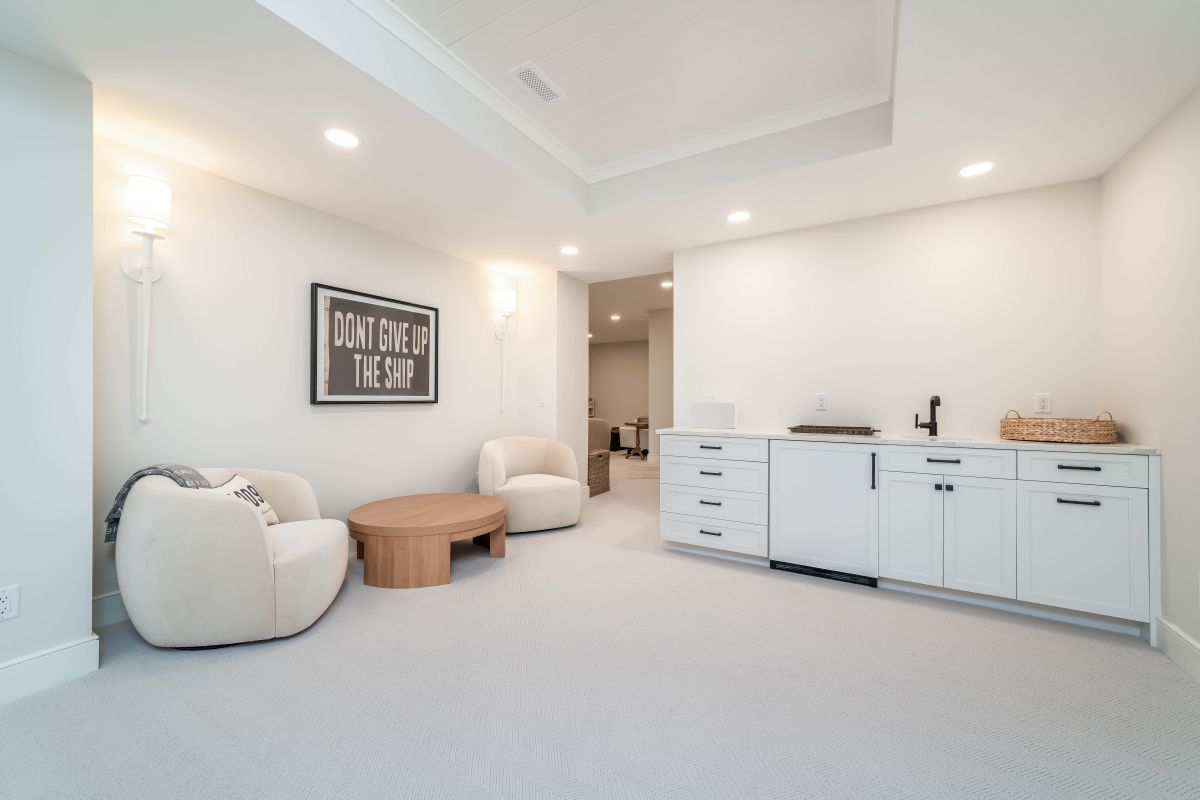 A bright basement living area features two cream armchairs, a round wooden coffee table, and a white wet bar with a sink.