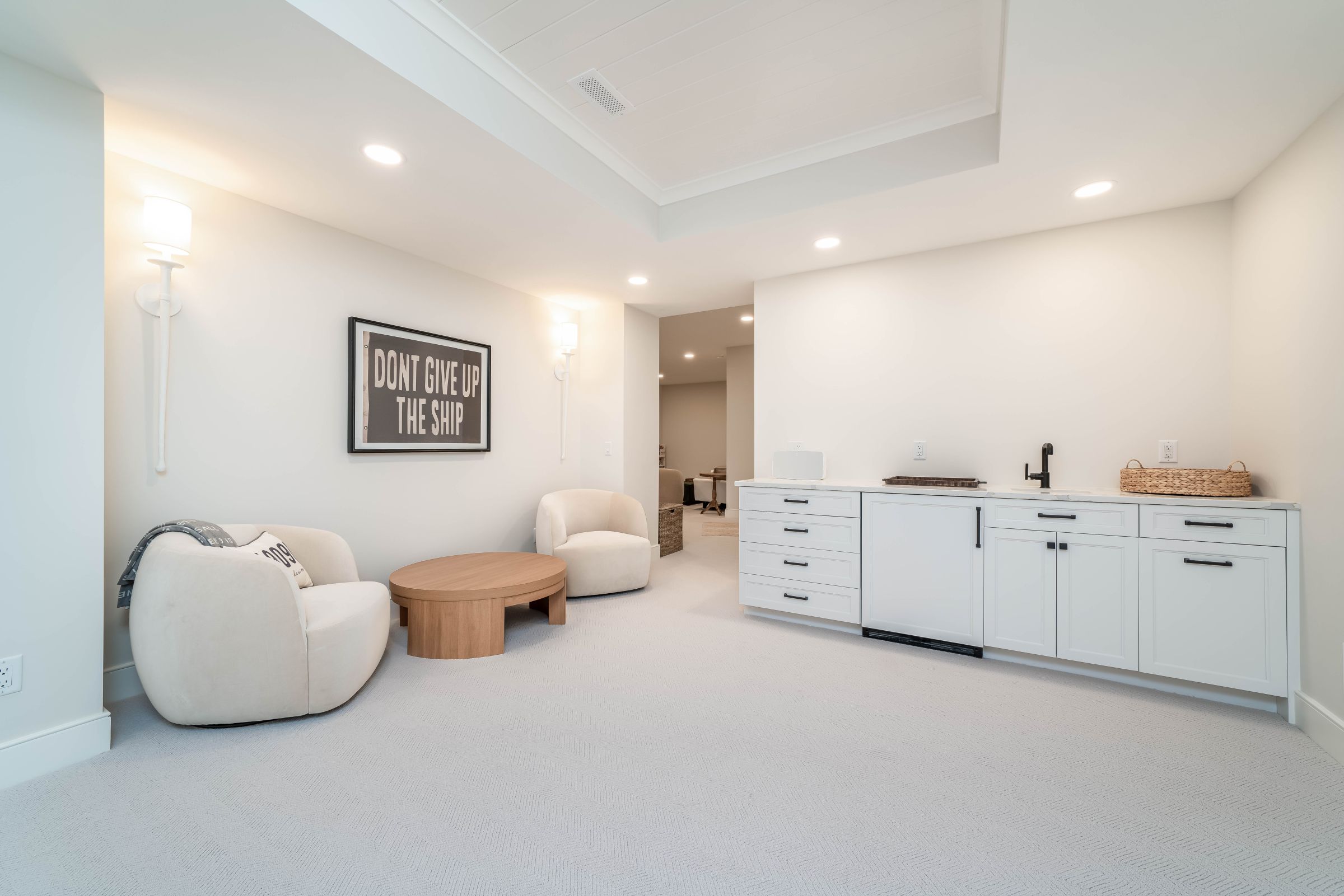 A bright basement living area features two cream armchairs, a round wooden coffee table, and a white wet bar with a sink.