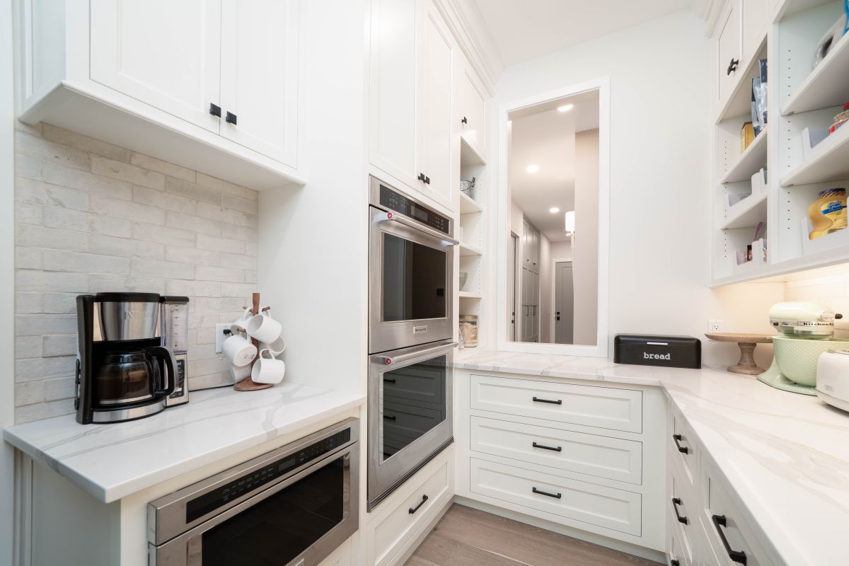 A white kitchen features a coffee maker, built-in ovens, a pantry with open shelving, and a window looking into a hallway.