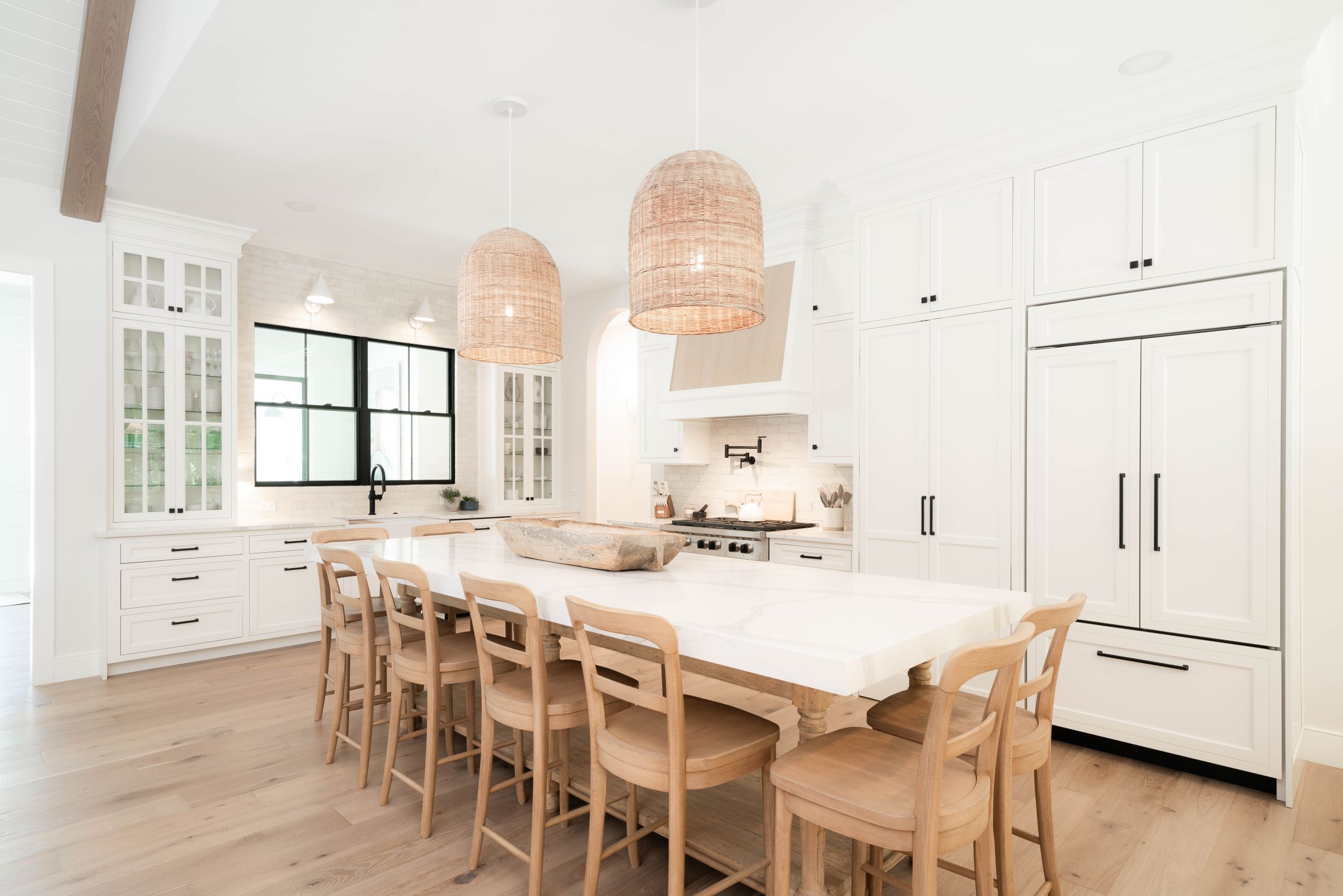 A bright white kitchen with a large island, light wood flooring, white cabinetry, a black-framed window, and two woven pendant lights.