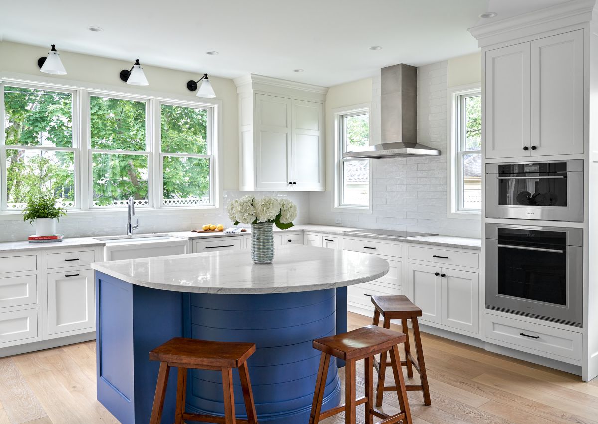 A bright kitchen features white shaker cabinets, light wood floors, a blue island with a marble top, and stainless steel appliances. Large windows overlook green trees.