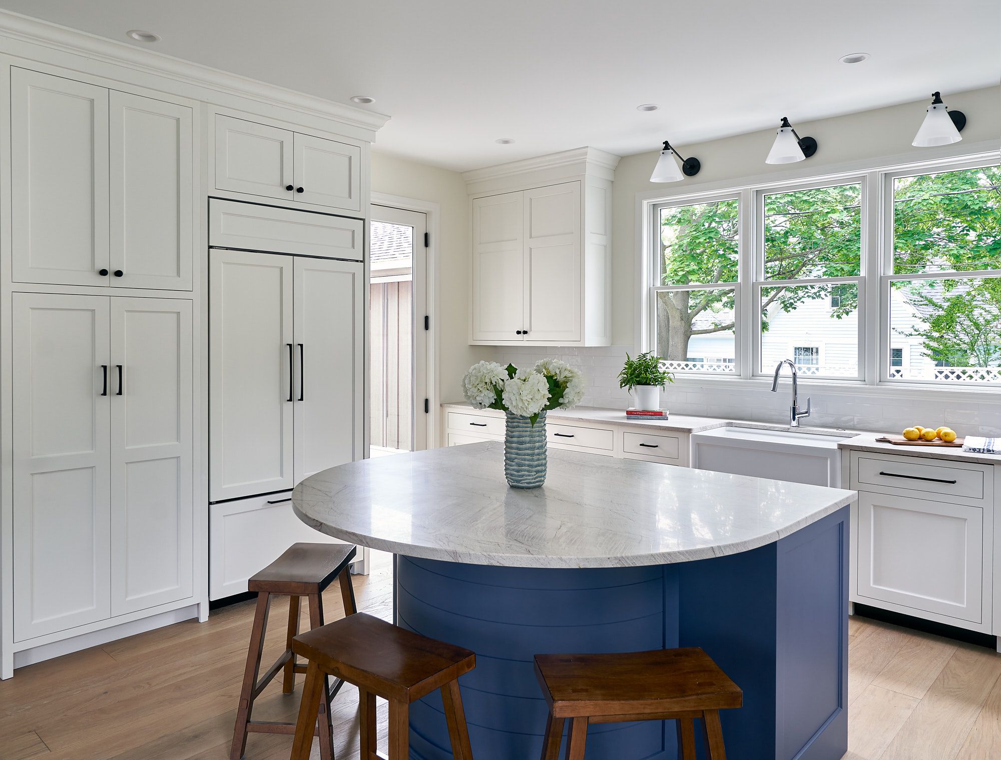 A white kitchen with a blue island, white cabinets, a large window over the sink, and wooden stools.