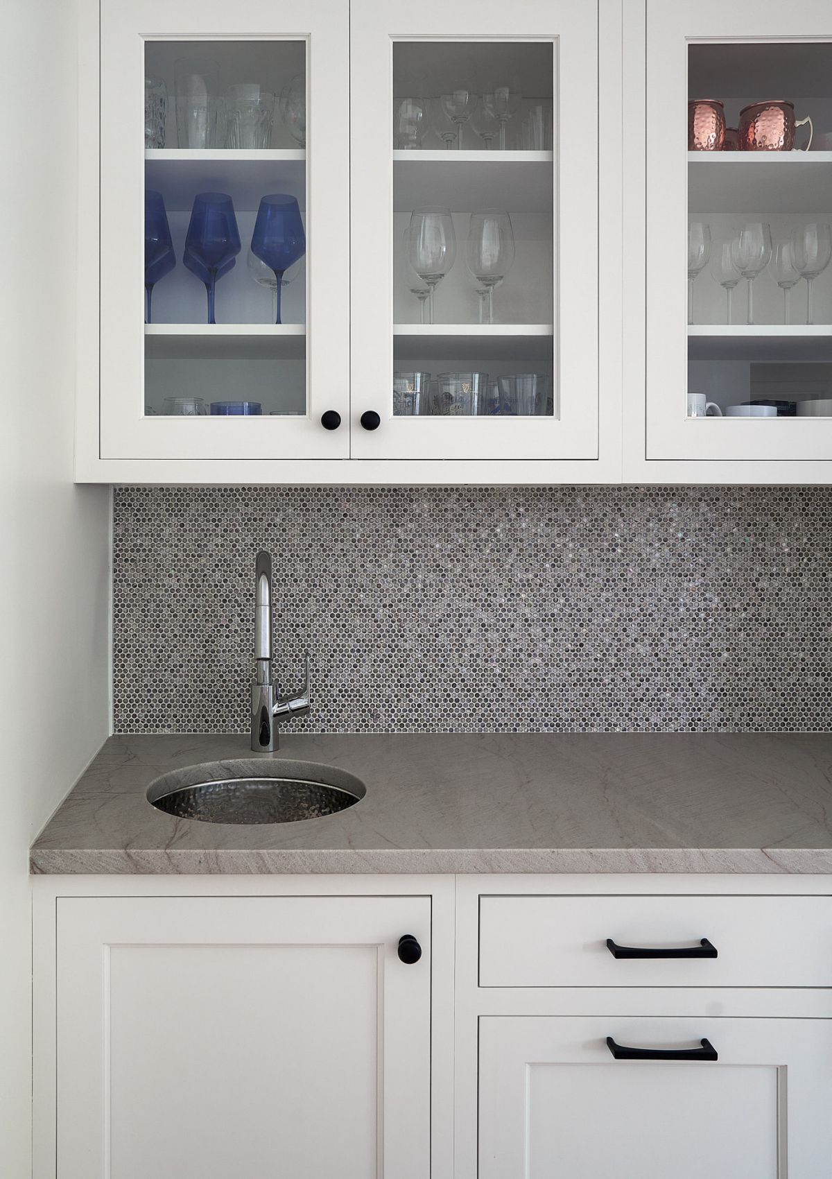 A white kitchen with glass-front upper cabinets, a gray countertop with a circular sink, and a gray mosaic tile backsplash.