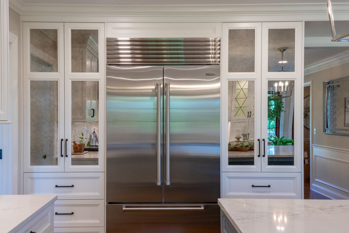 A kitchen with a built-in stainless steel refrigerator flanked by white cabinets with mirrored doors and a white countertop in the foreground.
