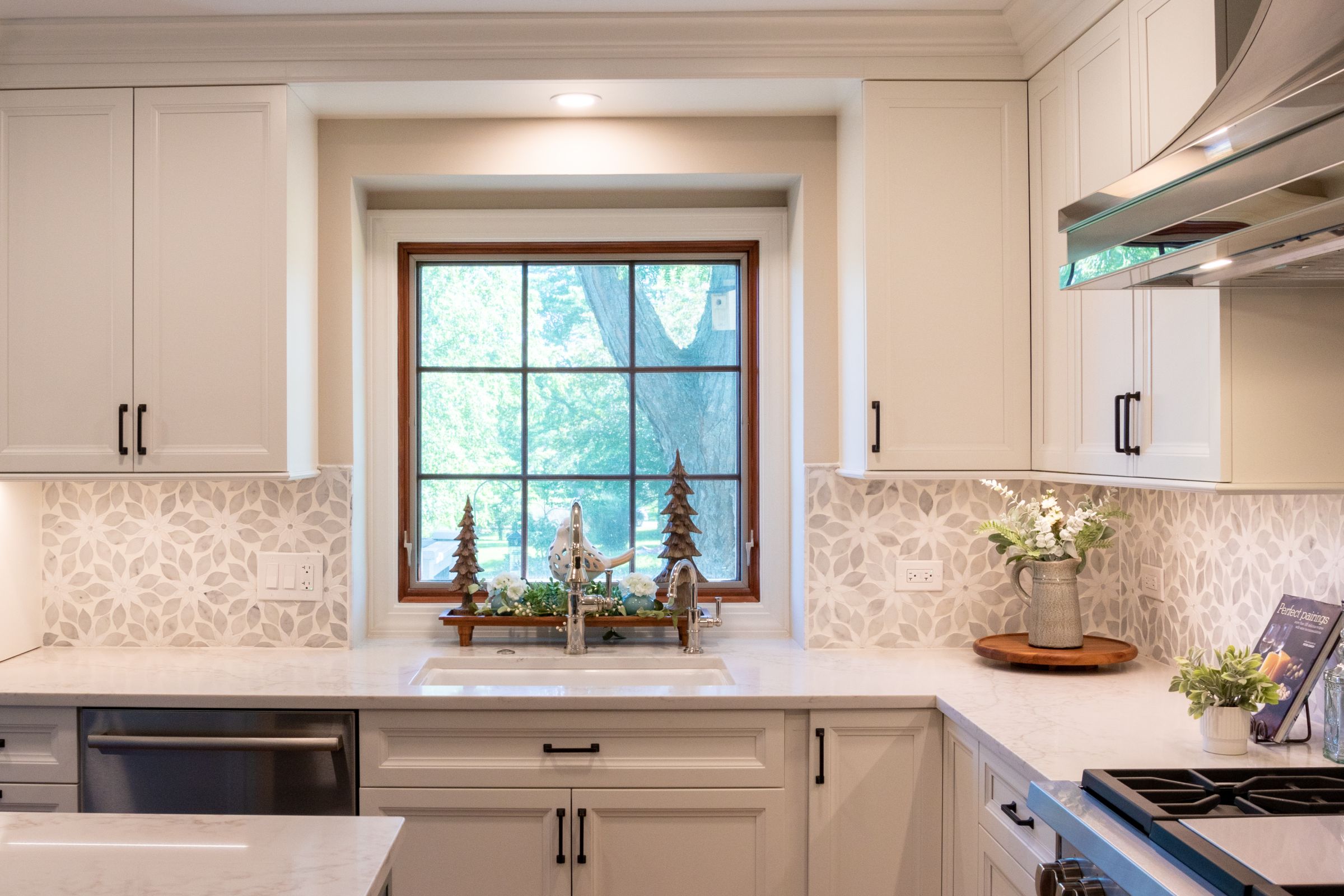 A kitchen features white upper and lower cabinets, a white countertop, a patterned backsplash, and a window above the sink.