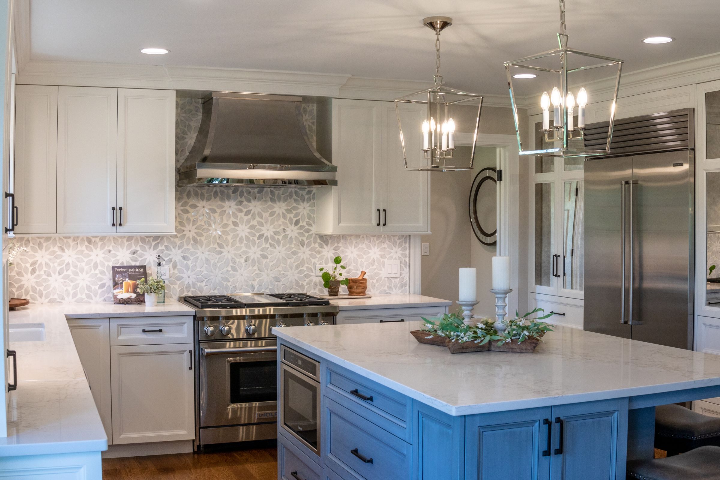 A kitchen with white upper and lower cabinets, a stainless steel range and refrigerator, a white and gray patterned backsplash, and a blue kitchen island with a white countertop.