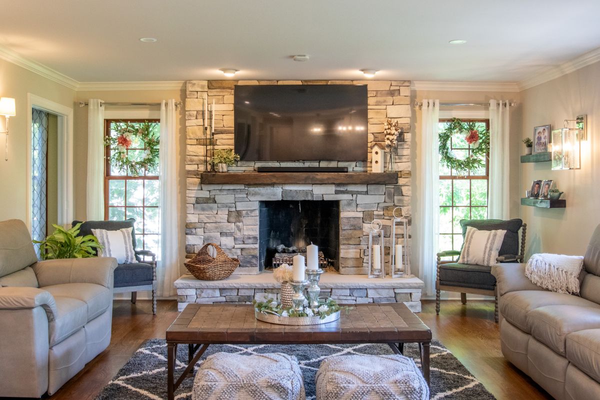 A living room with a stone fireplace, a large TV, two light-colored sofas, a dark wood coffee table, and two windows with wreaths.