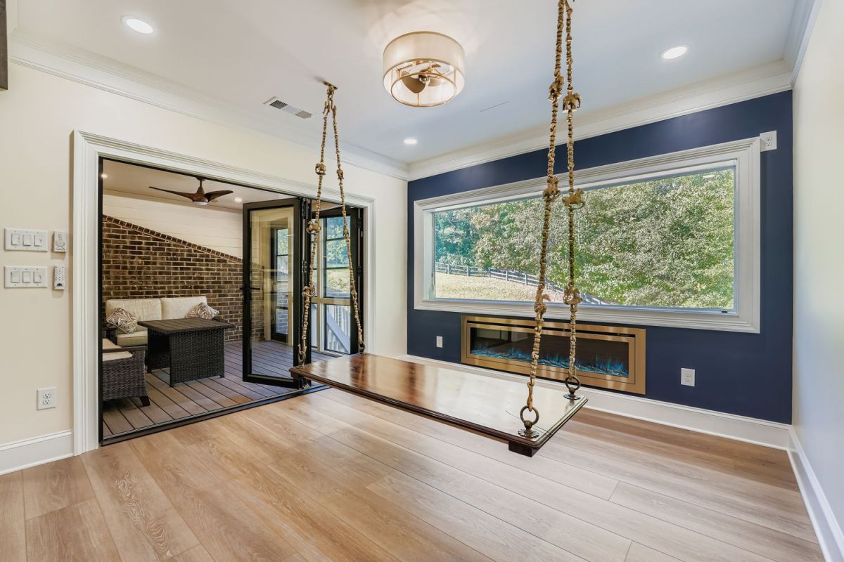 A room with light wood floors features a dark blue accent wall, a large window, a fireplace, and a wooden swing, with an outdoor patio visible through glass doors.