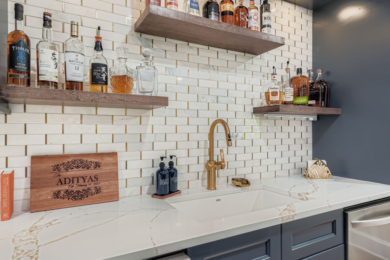 A basement bar with white subway tile, a gold faucet, a white sink, and dark wood shelves holding liquor bottles.