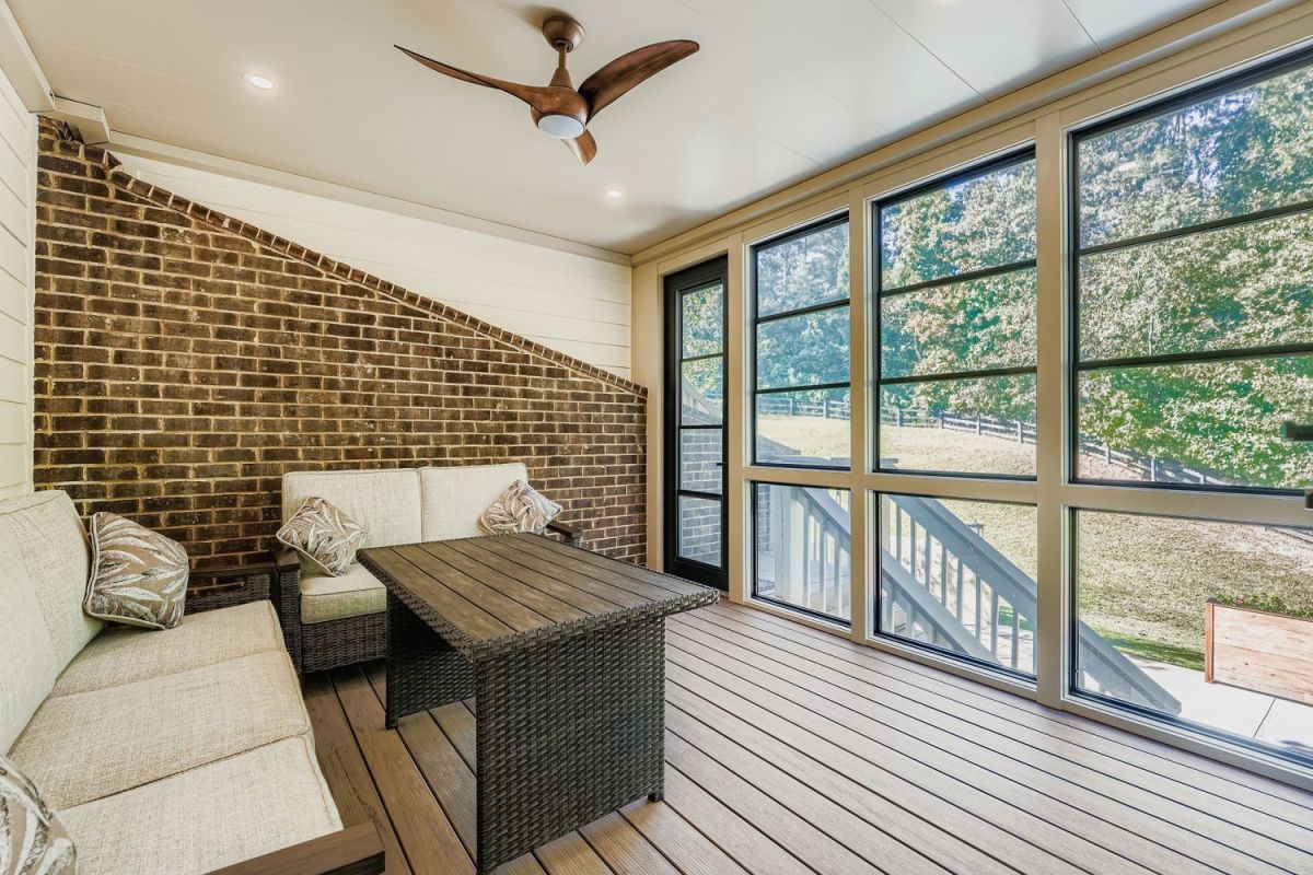 A screened-in porch with a brick accent wall, outdoor seating, a table, and large windows overlooking a grassy yard.
