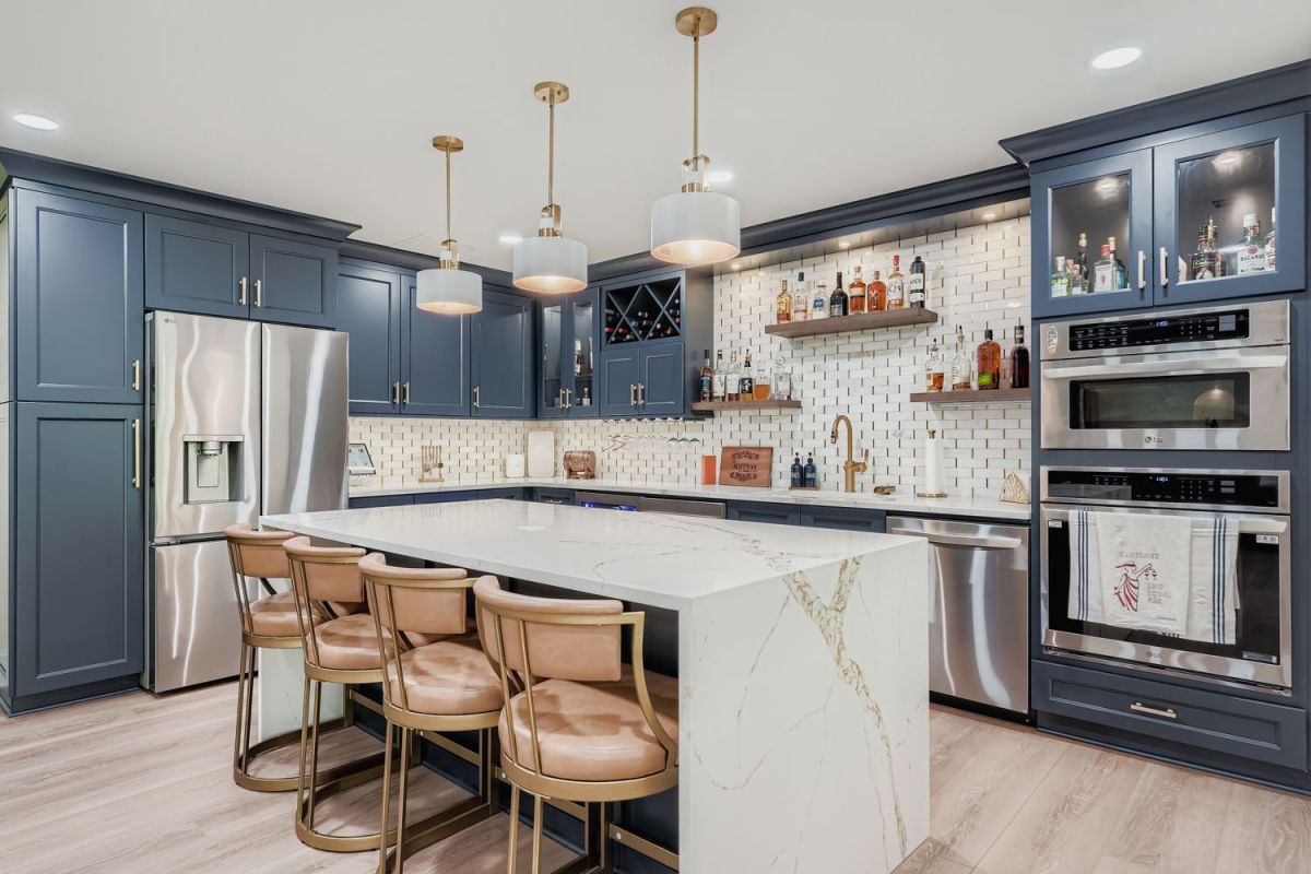 A basement kitchen and bar area with dark blue cabinets, a large white island with seating, stainless steel appliances, and a white tiled backsplash with open shelving for bottles.