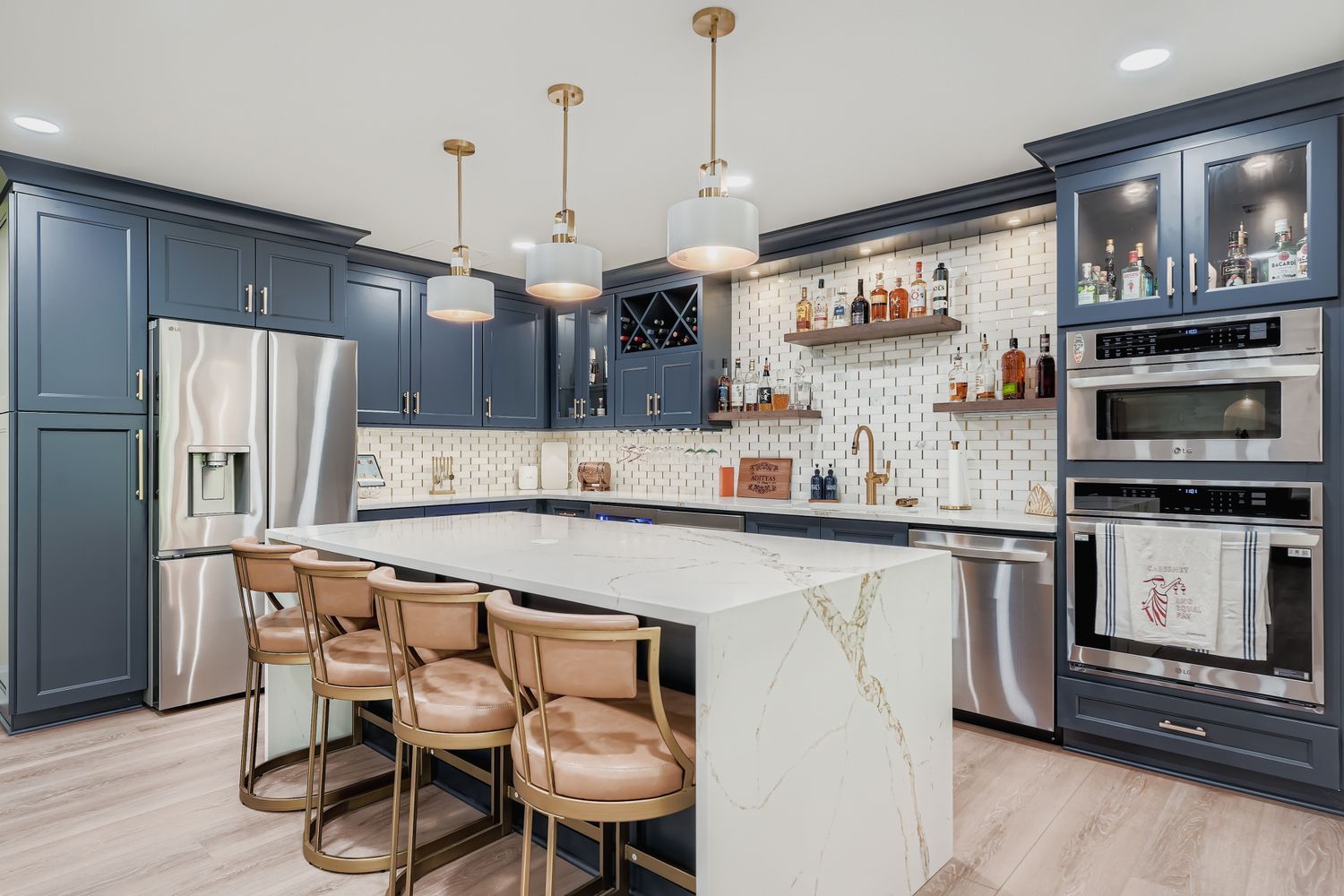 A basement kitchen and bar area with dark blue cabinets, a large white island with seating, stainless steel appliances, and a white tiled backsplash with open shelving for bottles.