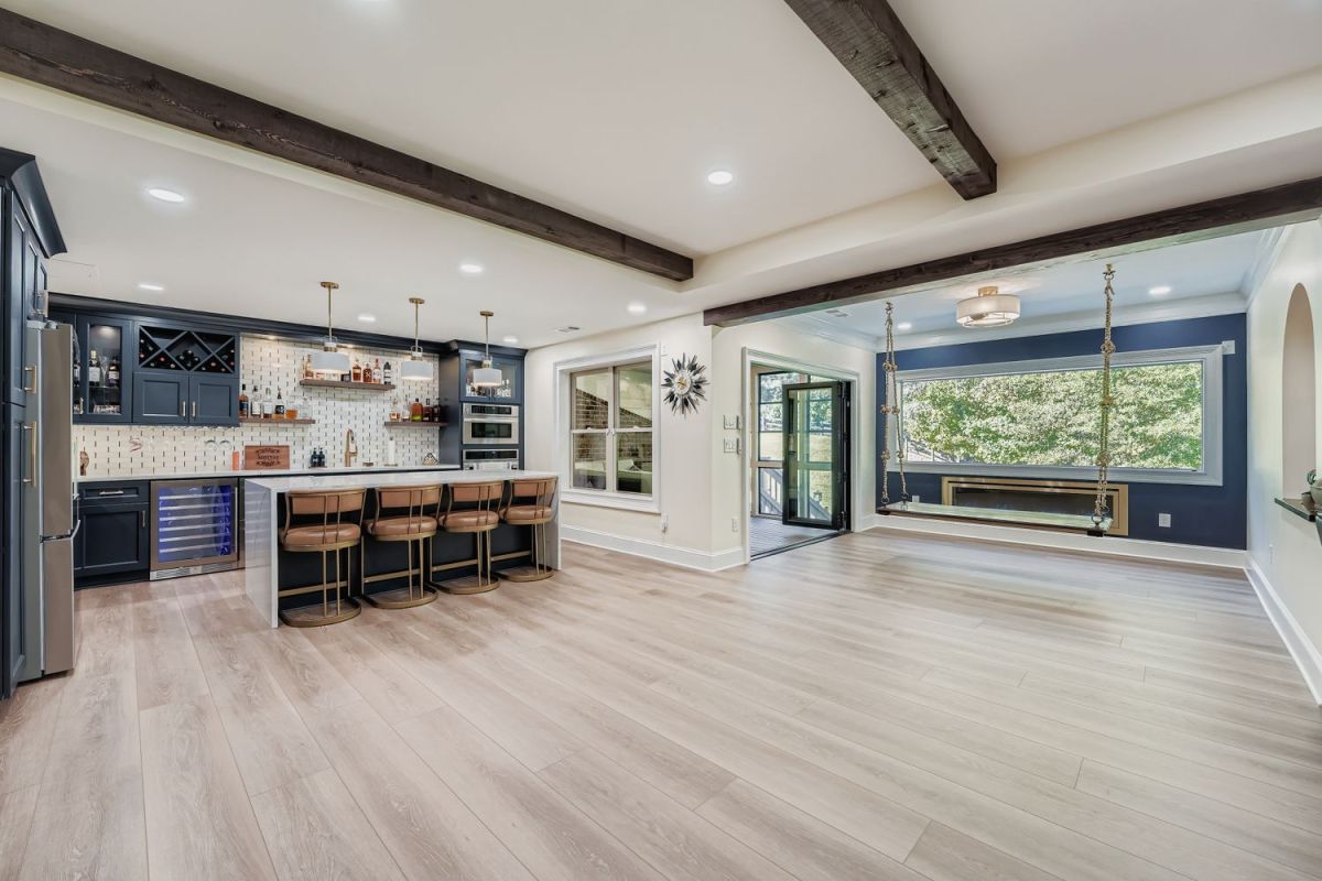 A wide-angle view of a finished basement featuring a dark blue bar with a white countertop and four bar stools, light wood flooring, and a lounge area with a hanging swing.