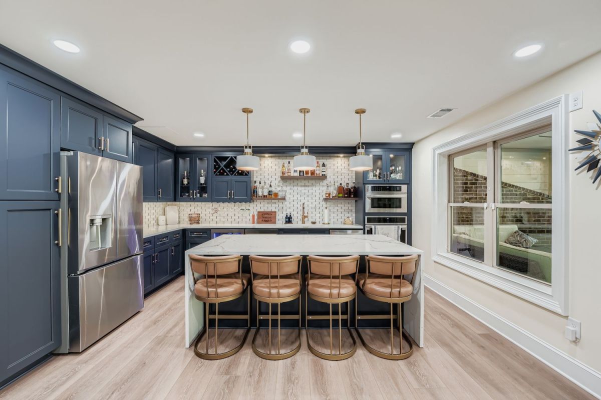 A basement bar features blue shaker cabinets, a large island with four bar stools, stainless steel appliances, and a window.