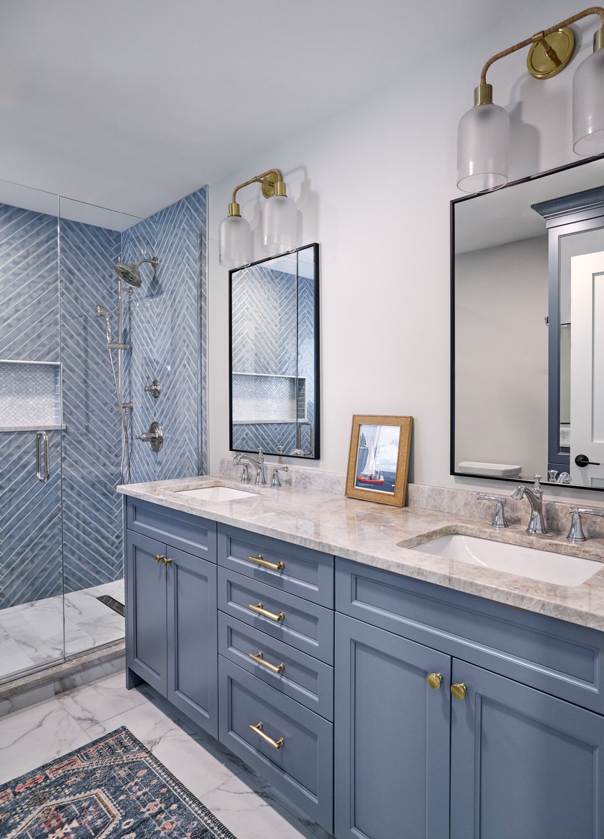 A bathroom features a double vanity with blue cabinets, a marble countertop, and two mirrors, alongside a glass-enclosed shower with blue herringbone tiles.