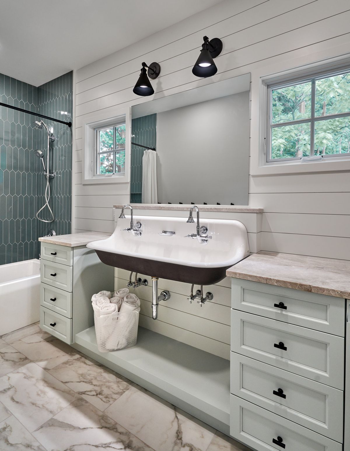 A bathroom features a double trough sink with two faucets, a large mirror, light green vanity cabinets, and a shower with green picket tiles.