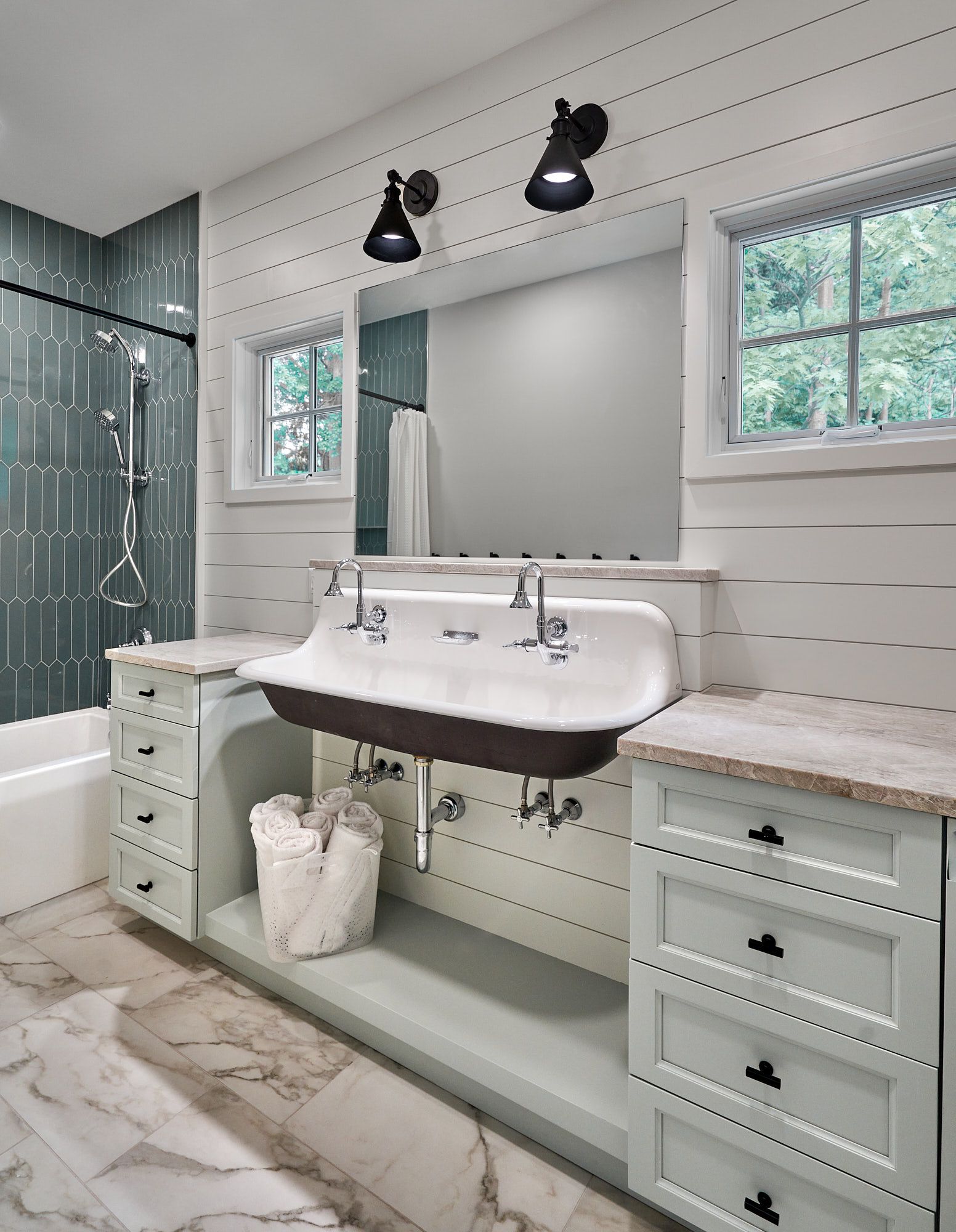 A bathroom features a double trough sink with two faucets, a large mirror, light green vanity cabinets, and a shower with green picket tiles.