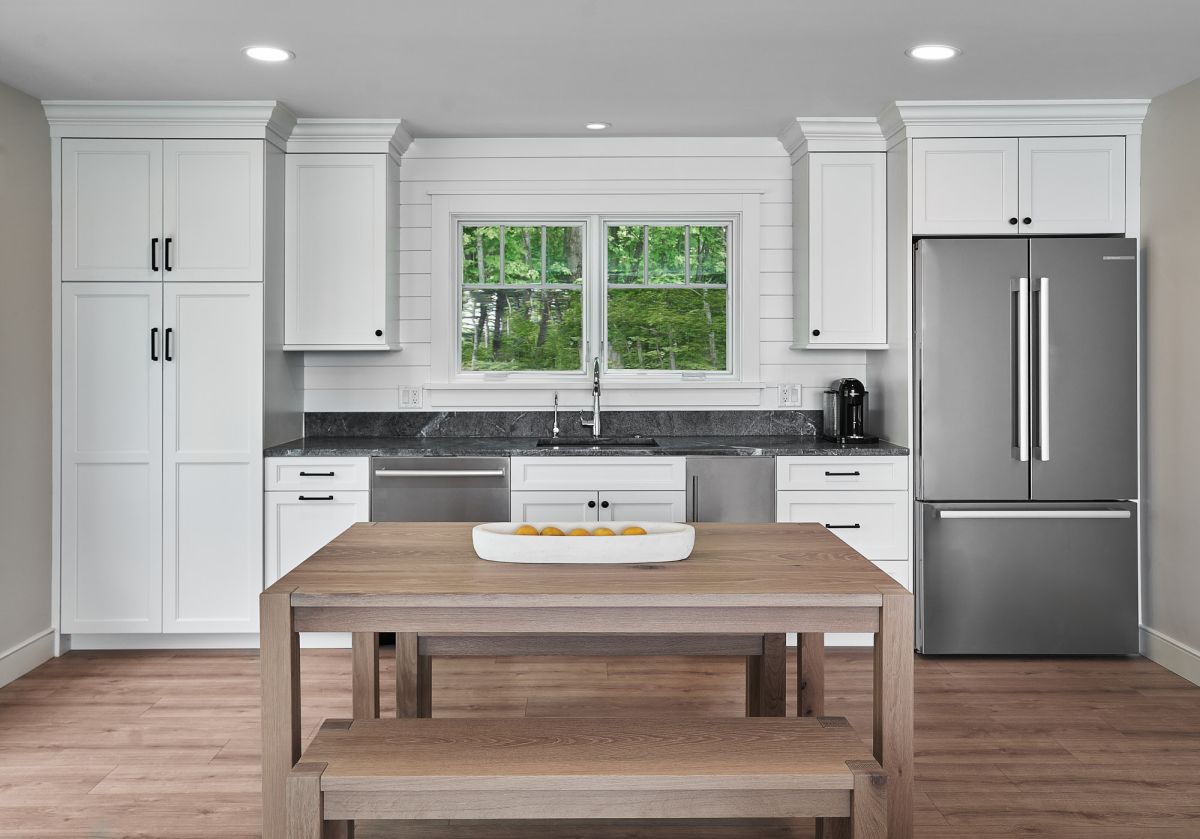 A kitchen with white shaker cabinets, dark countertops, a stainless steel refrigerator, and a wooden dining table with a bench.