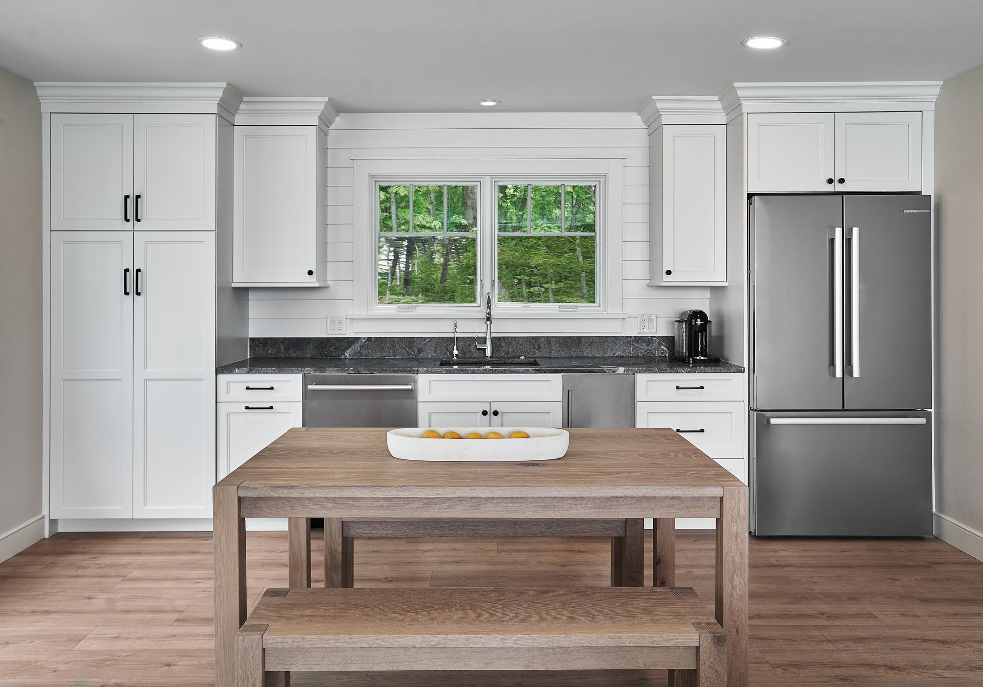 A kitchen with white shaker cabinets, dark countertops, a stainless steel refrigerator, and a wooden dining table with a bench.