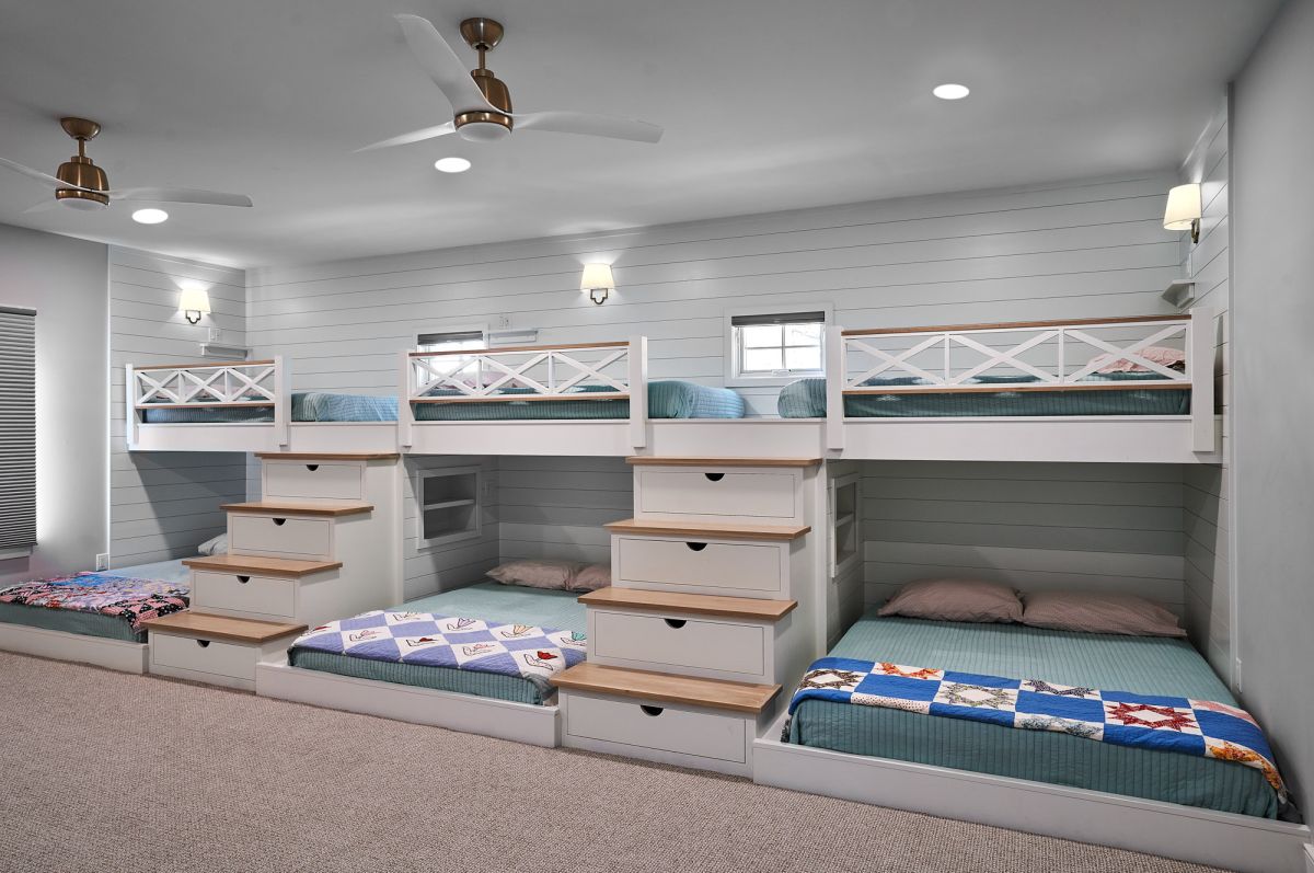 A large bedroom with four built-in bunk beds, two on each side, featuring white frames, wood accents, and light blue bedding. Two ceiling fans and wall sconces provide lighting.