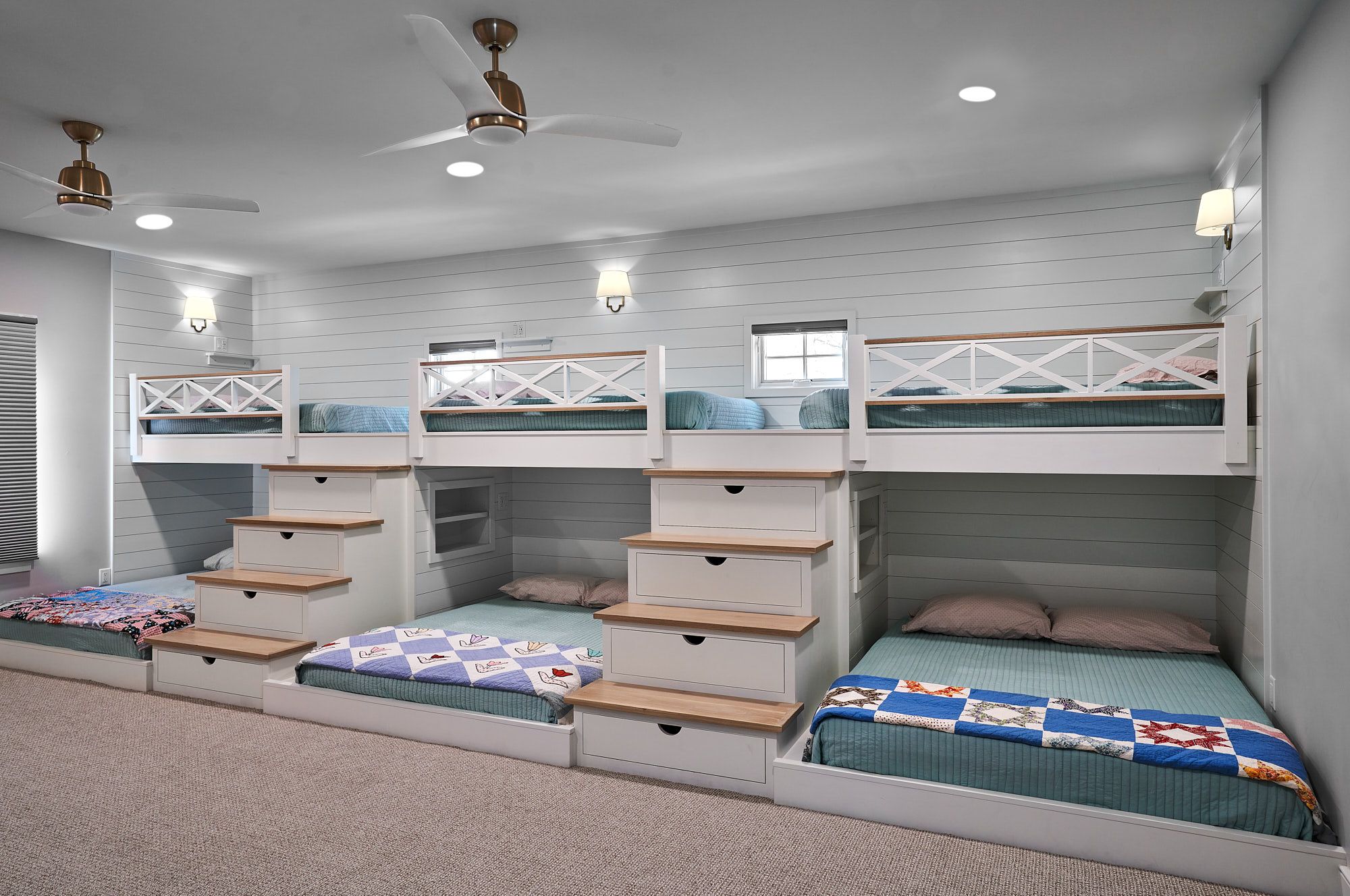 A large bedroom with four built-in bunk beds, two on each side, featuring white frames, wood accents, and light blue bedding. Two ceiling fans and wall sconces provide lighting.