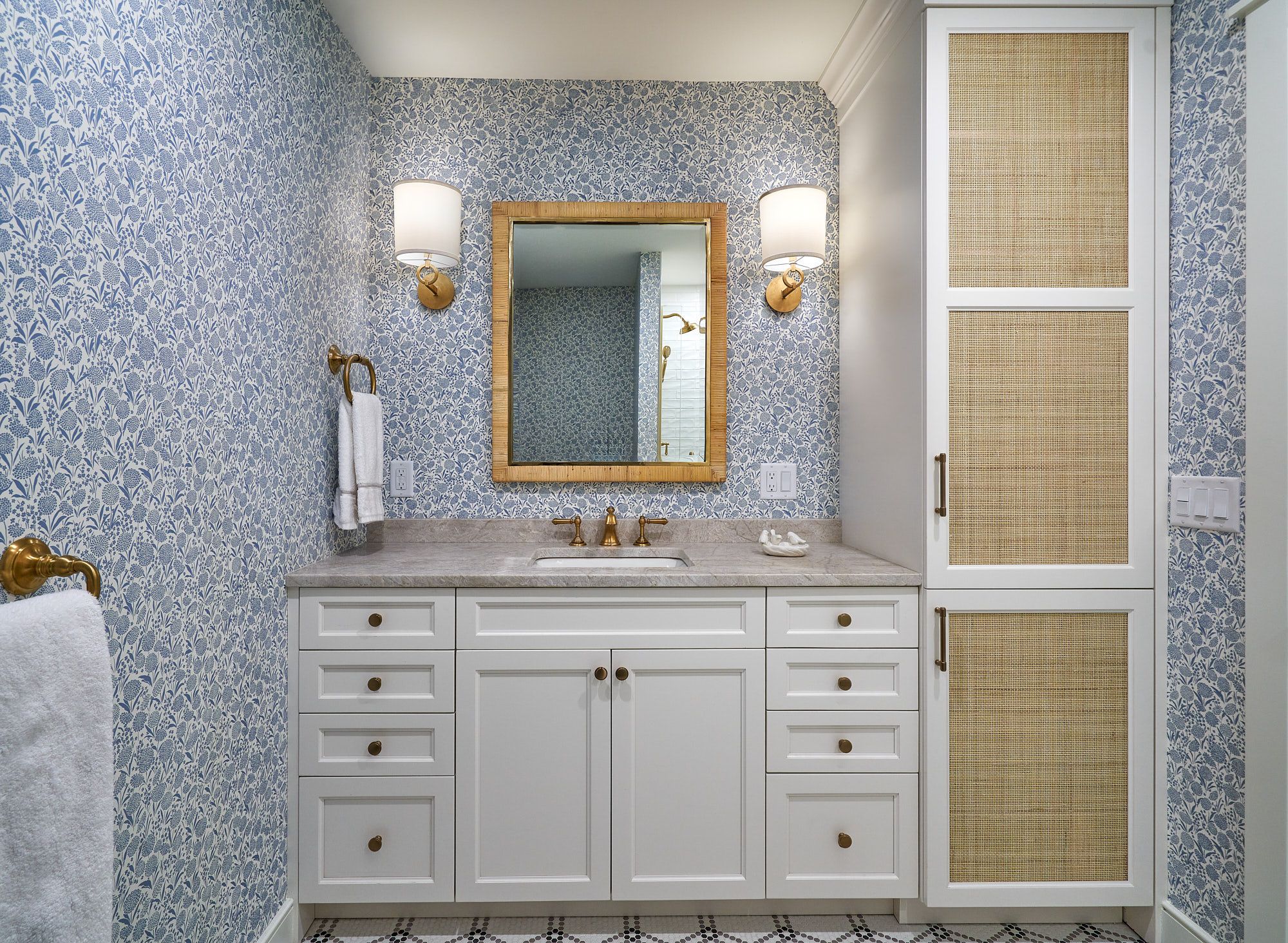 A bathroom features blue and white patterned wallpaper, a white vanity with a light-colored countertop, a mirror, and a tall storage cabinet.