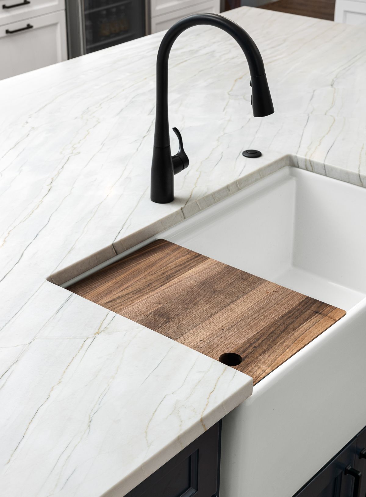 A kitchen island features a white marble countertop with a black faucet, a white farmhouse sink, and a wooden cutting board.