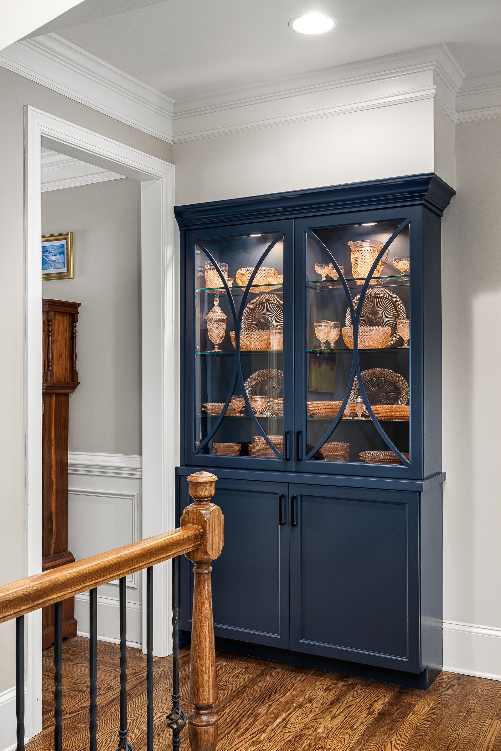 A navy blue display cabinet with glass doors and internal lighting stands on a hardwood floor, next to a white doorway and a wooden stair railing.