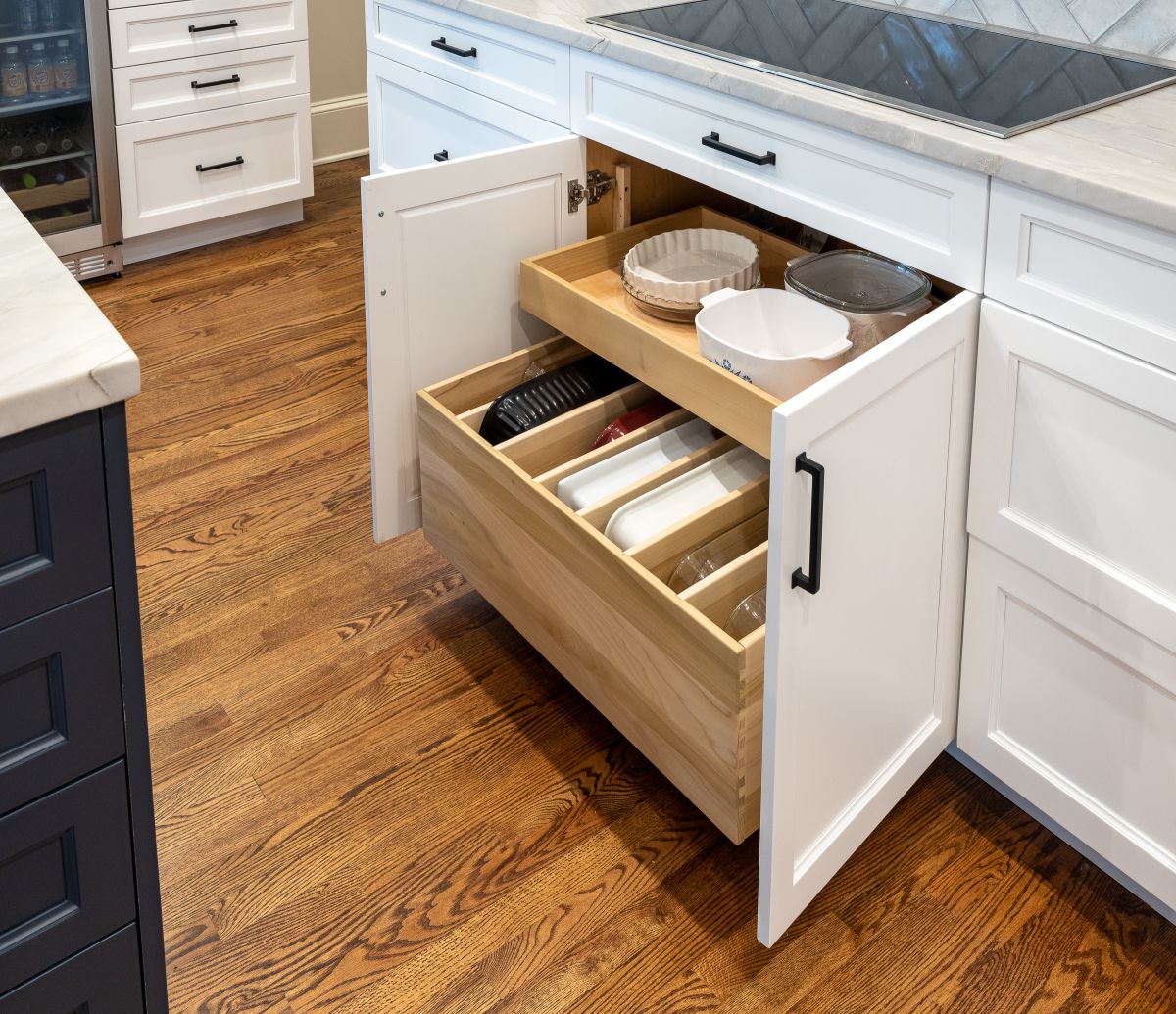 A kitchen with hardwood floors, white and dark blue cabinets, and a pull-out drawer system under a cooktop.