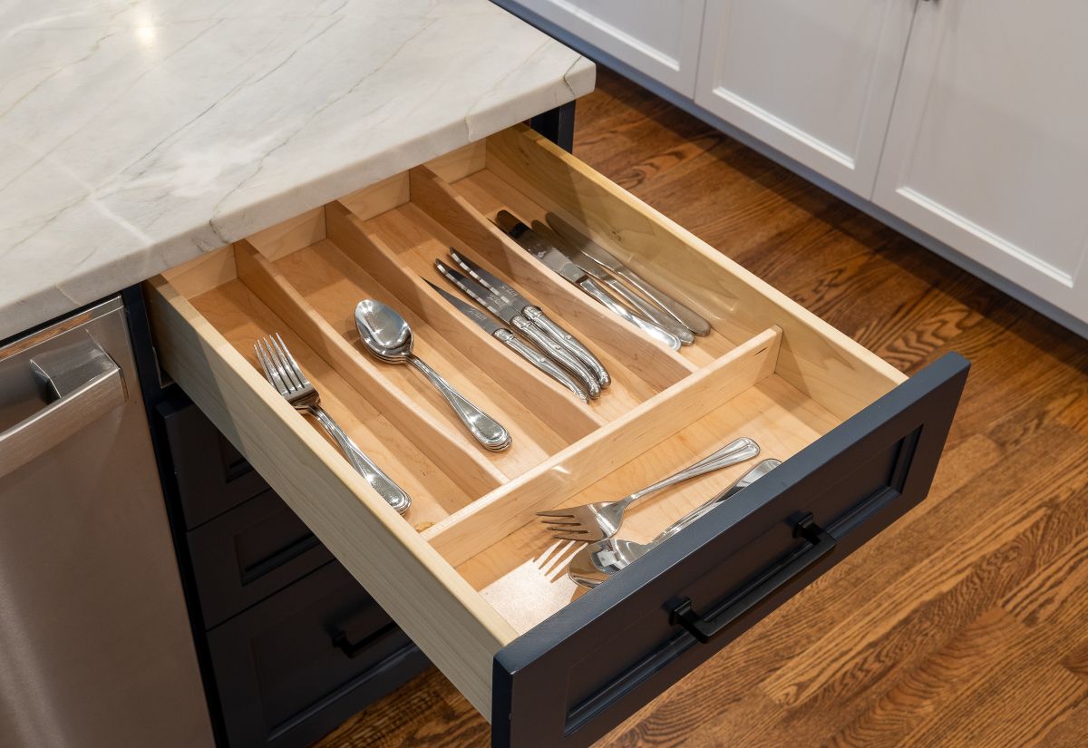 An open kitchen drawer with a wooden organizer holds various silverware, next to a stainless steel appliance and a marble countertop.