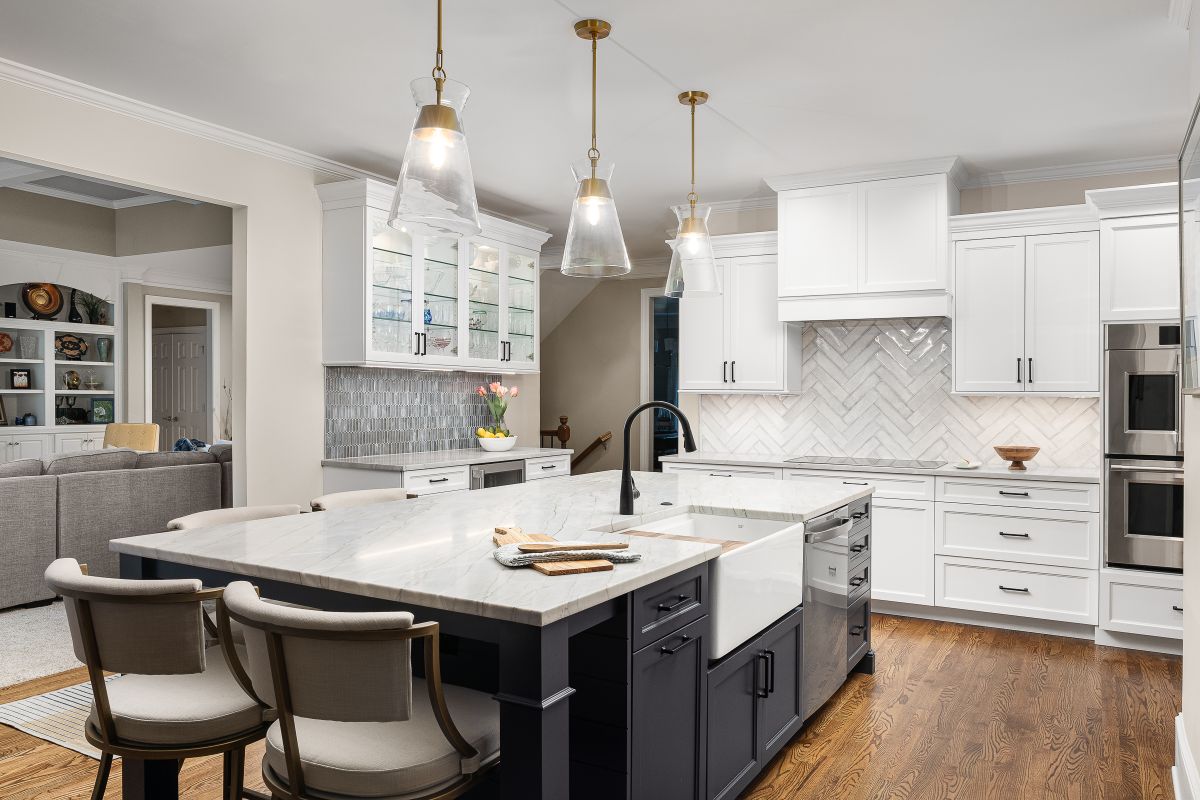 A white kitchen with a large island, dark gray base cabinets, and white upper cabinets, with an adjacent living room visible.